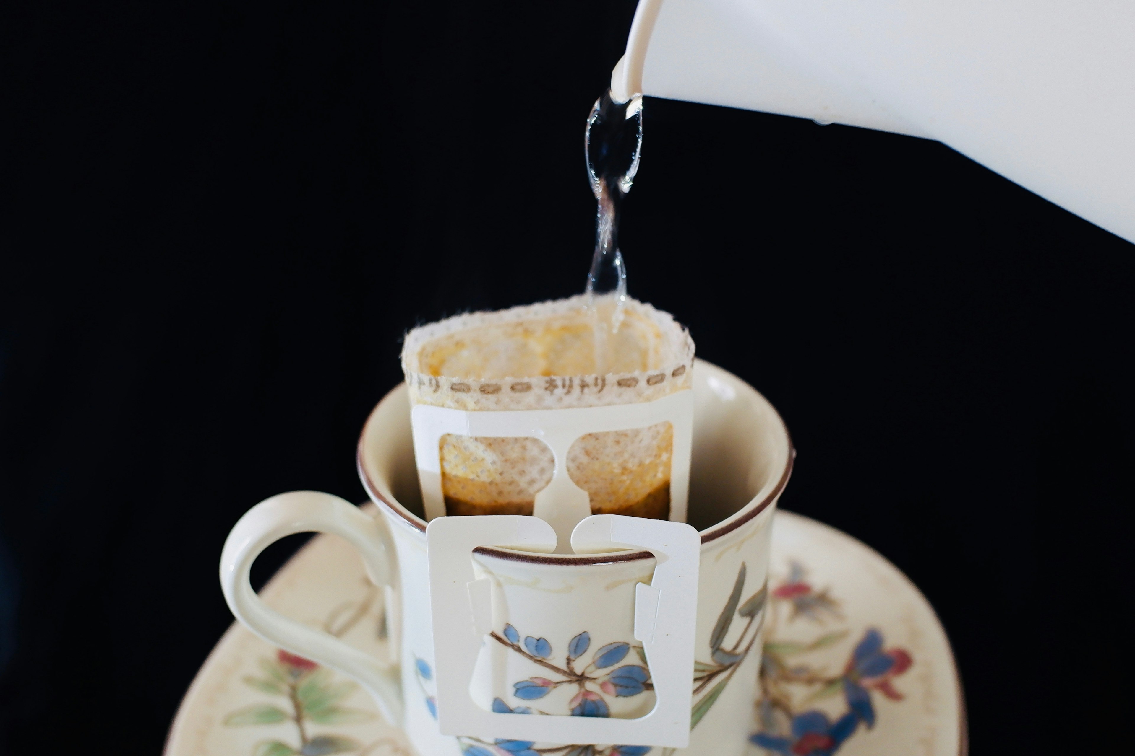 Tee wird aus einer weißen Teekanne in eine Tasse mit floralem Muster und Teebeutelhalter gegossen