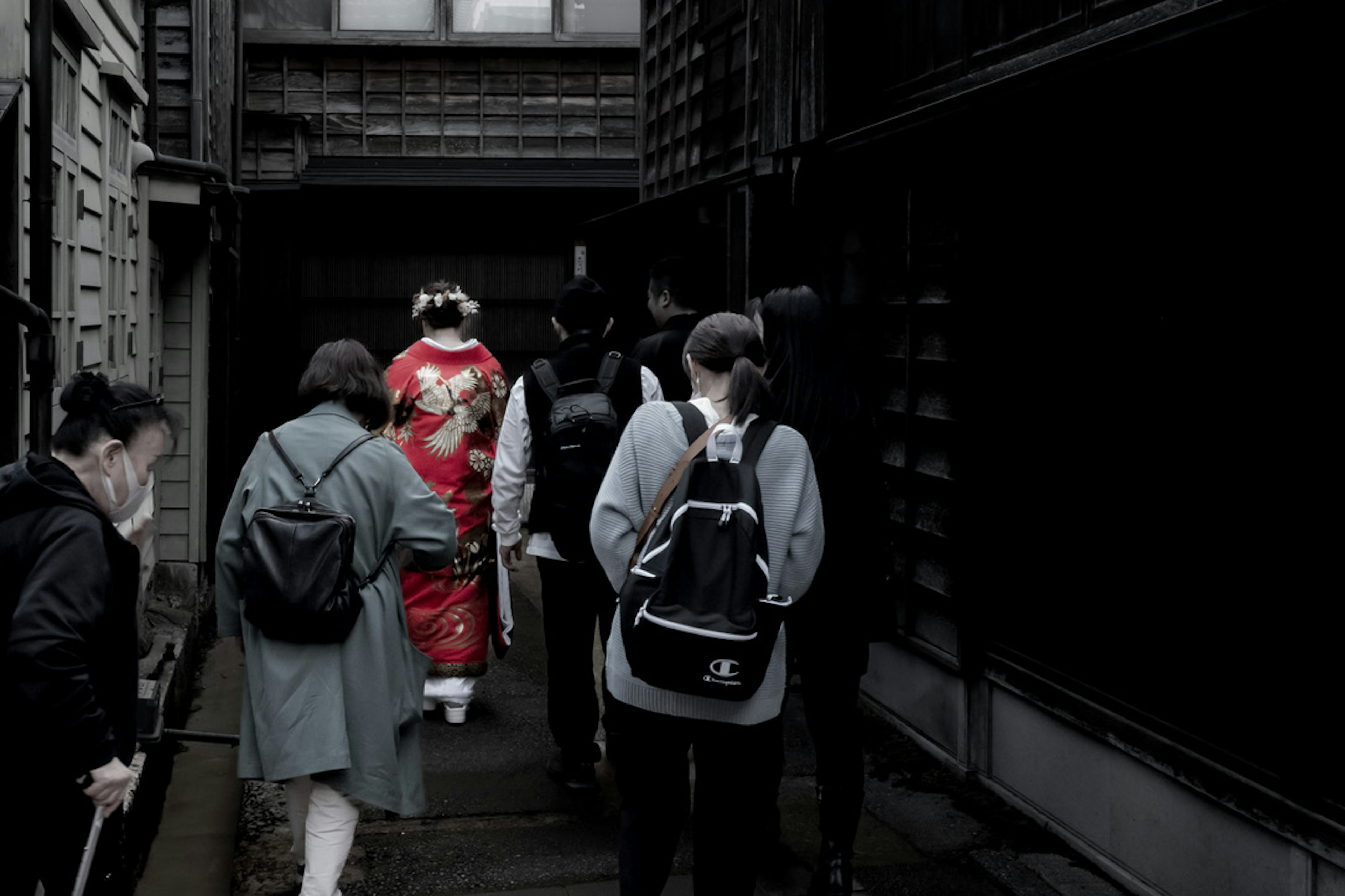 Group of people walking through a narrow passage behind a woman in a red kimono