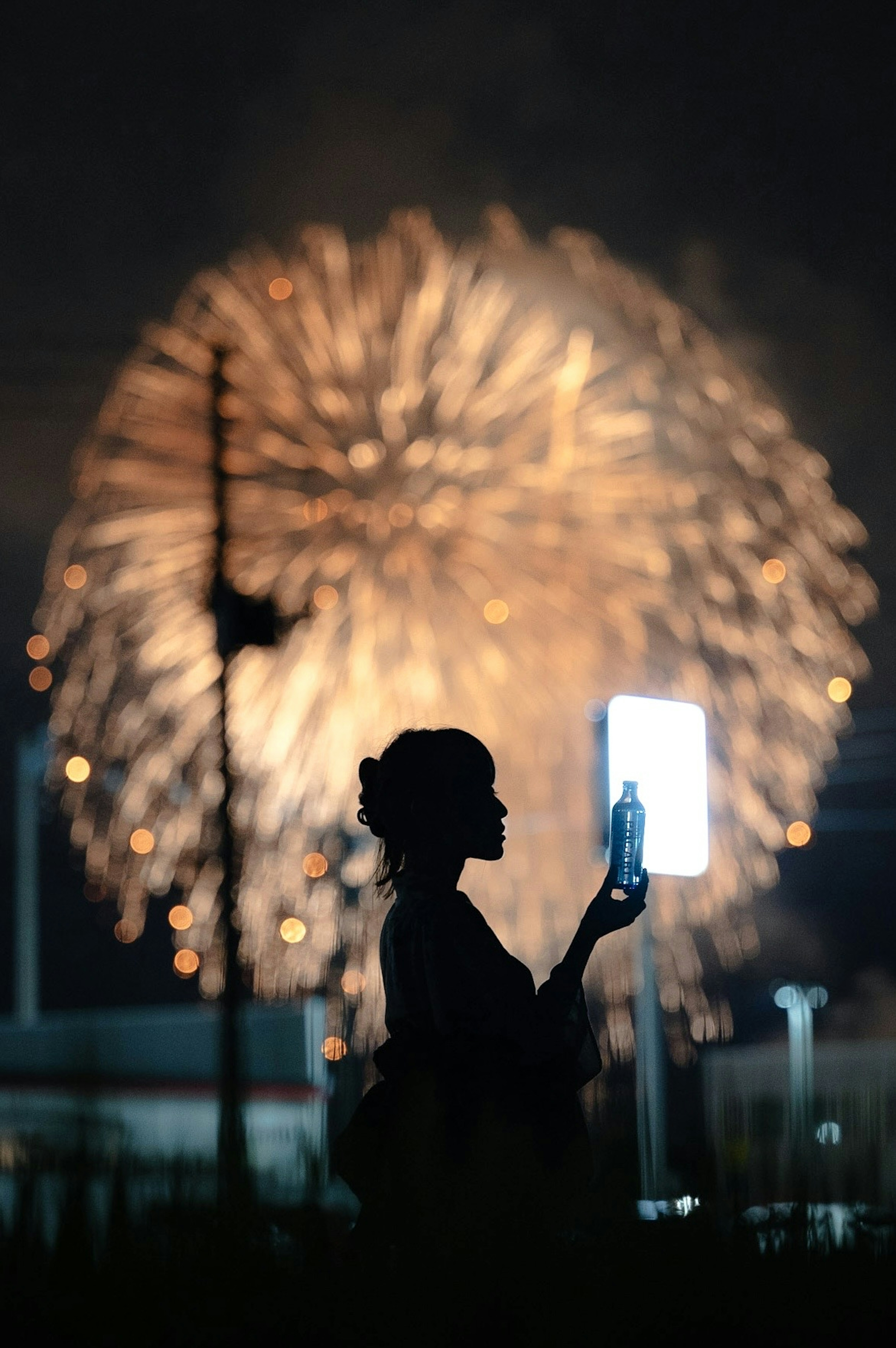 夜空に花火が打ち上がる中、着物を着た女性がスマートフォンを持っているシルエット