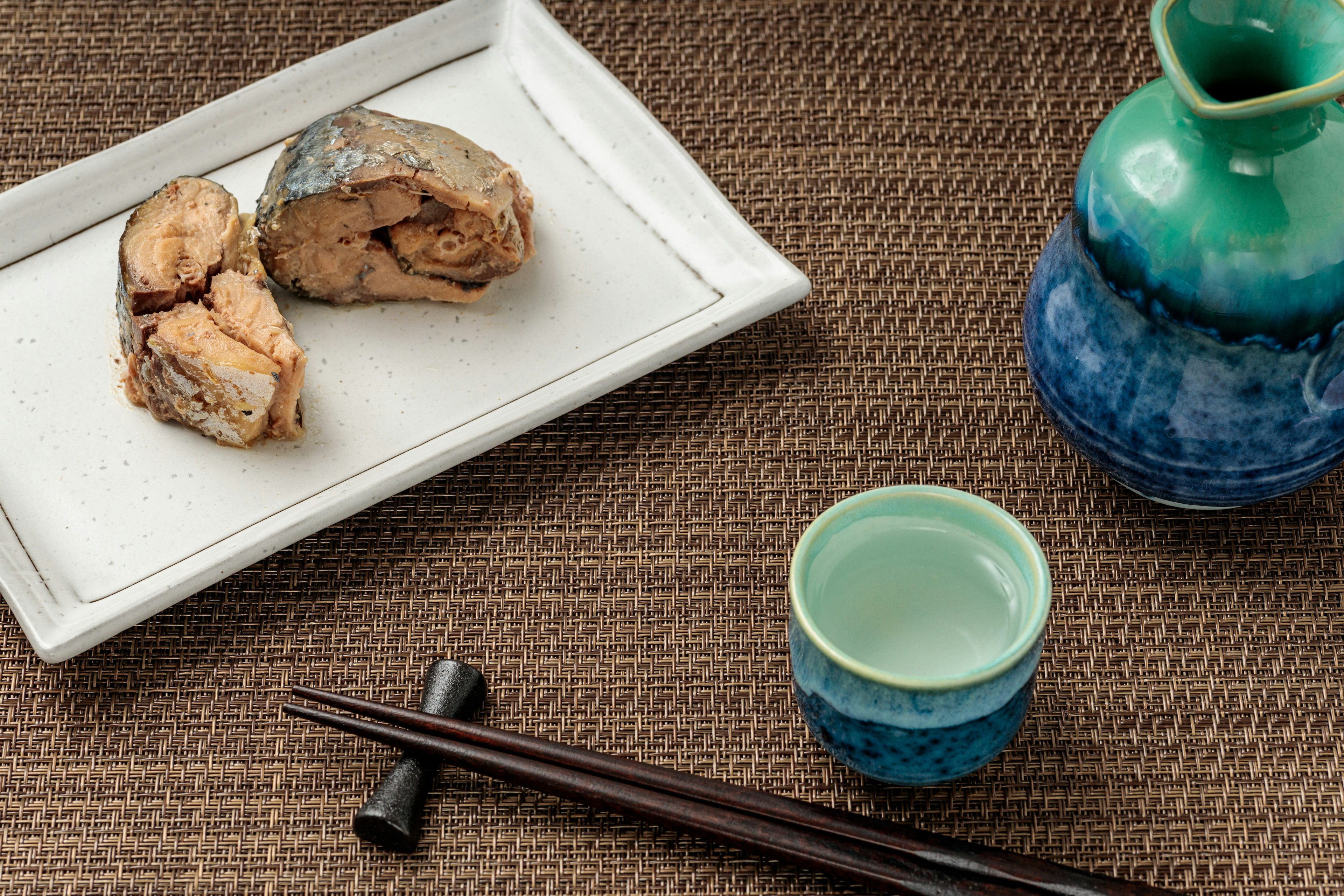 Ikan bakar di piring putih dengan botol sake biru dan cangkir kecil di atas meja bertekstur