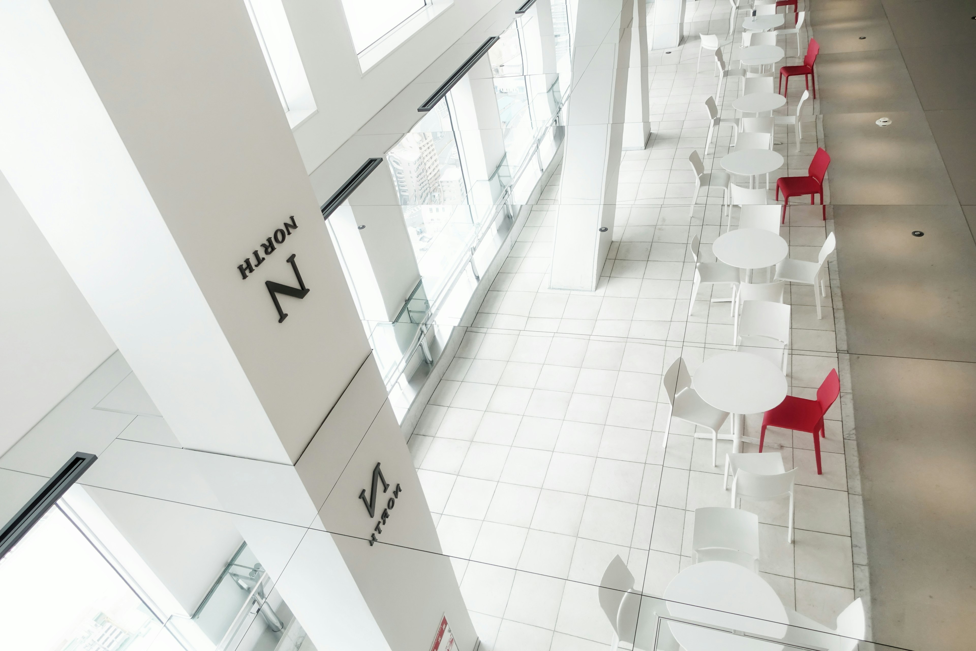 Modern café area with white walls and large windows featuring red chairs and white tables