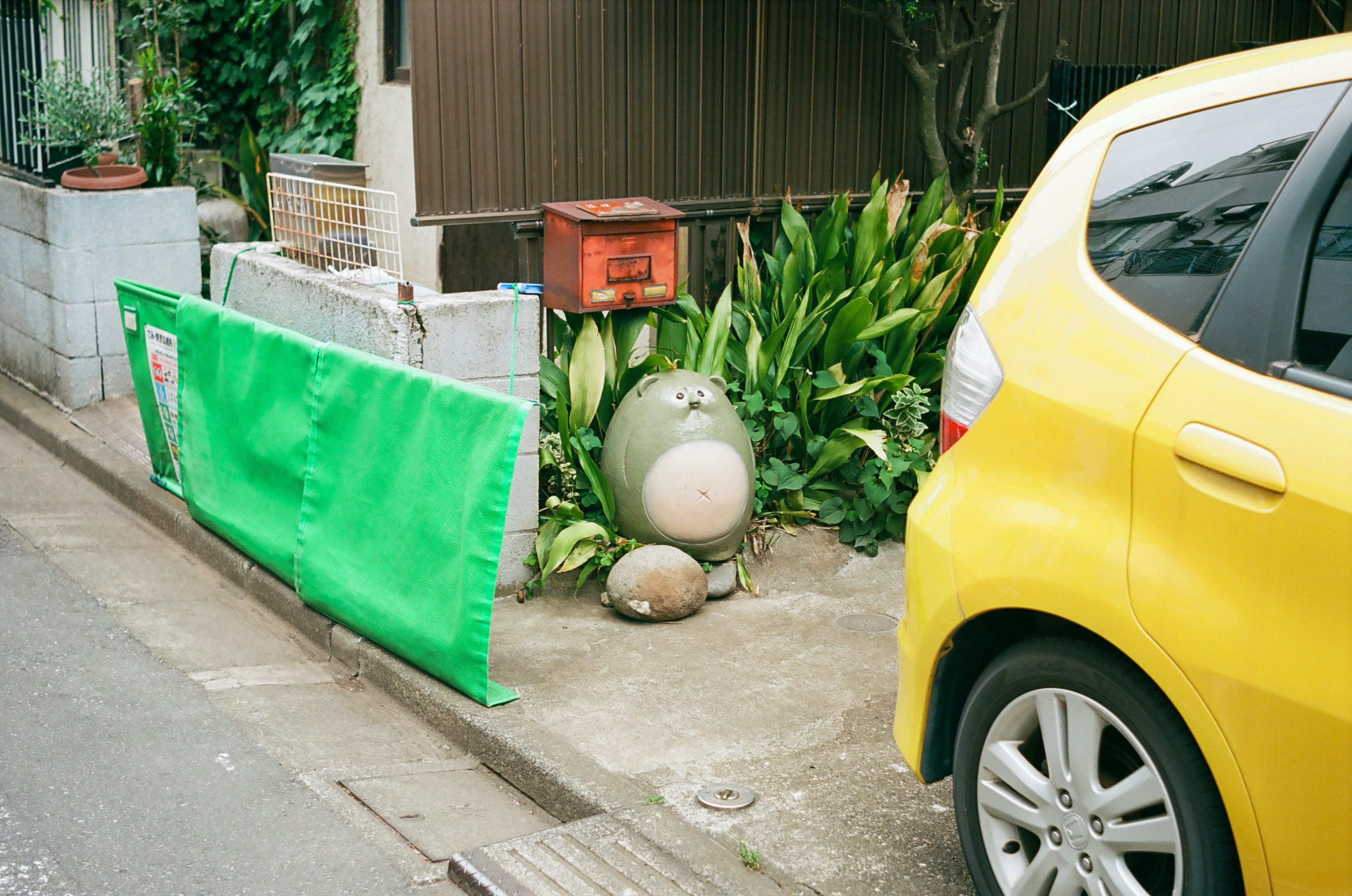 Una scena di strada con un'auto gialla e una copertura verde, piante e una cassetta delle lettere