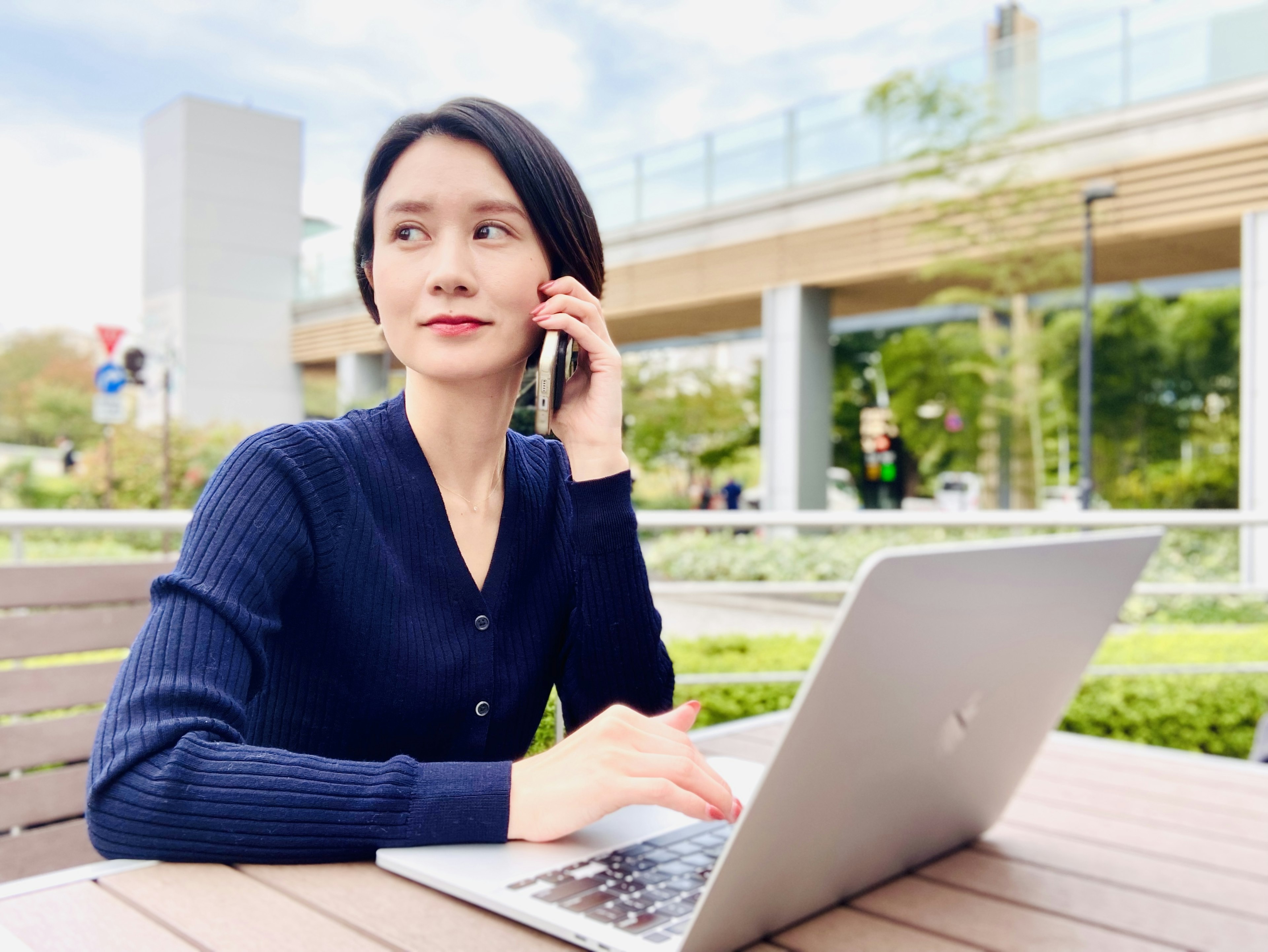 Eine Frau sitzt auf einer Parkbank und benutzt einen Laptop, während sie telefoniert