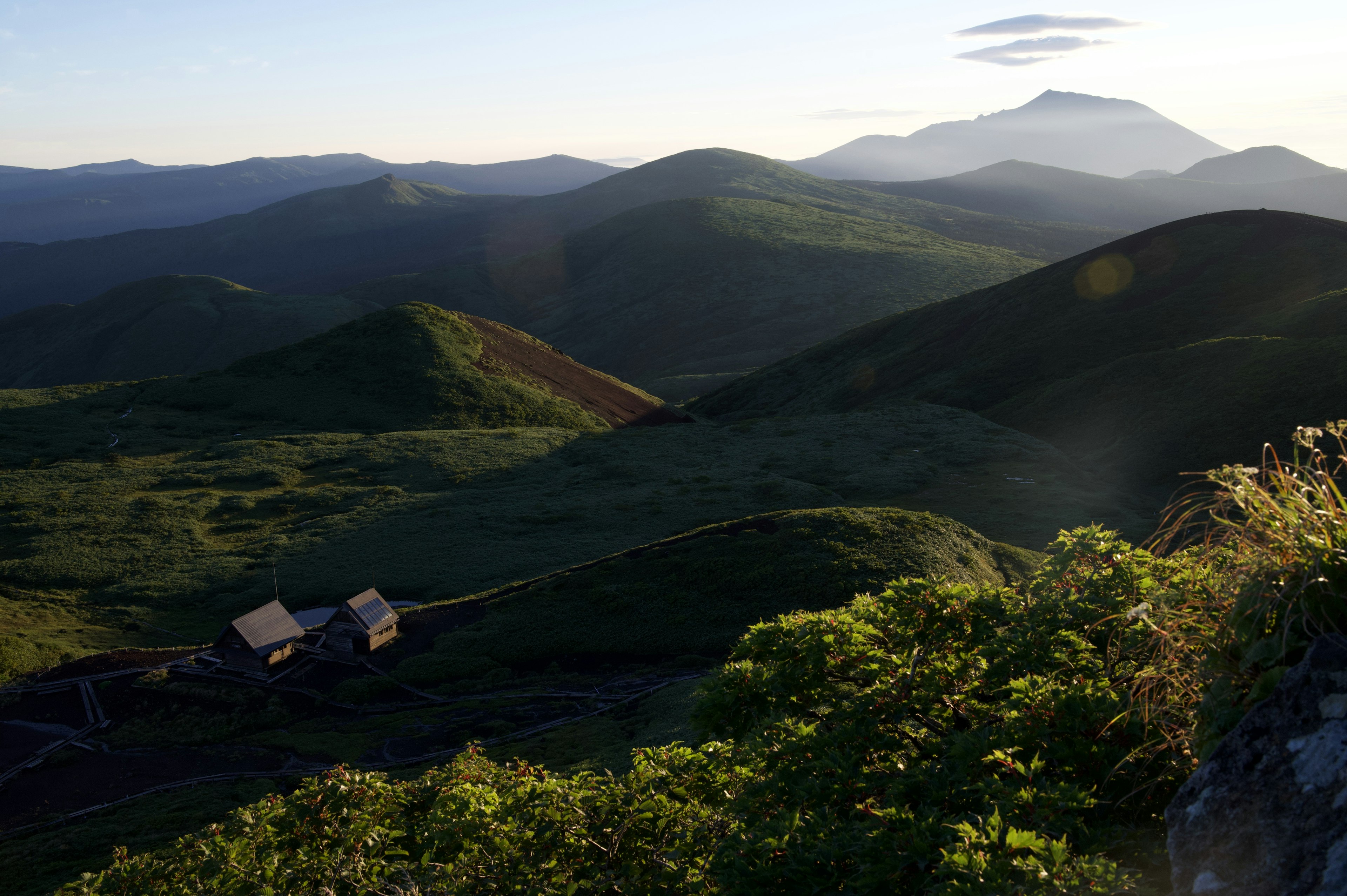 緑の丘陵と遠くの山々を背景にした風景、夕暮れ時の柔らかな光が差し込む