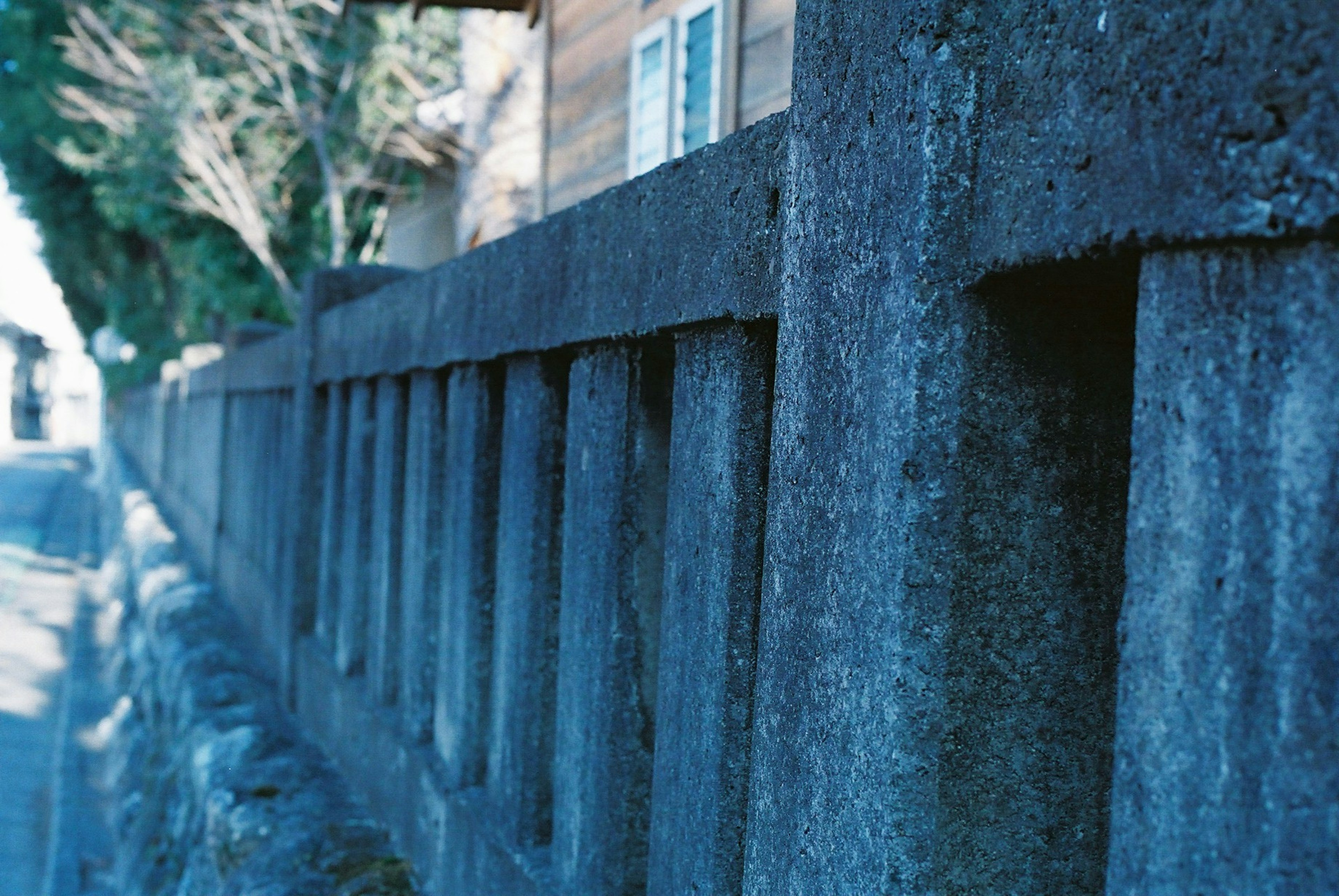 Close-up pagar beton dengan nada kebiruan, latar belakang terdapat pohon dan bangunan