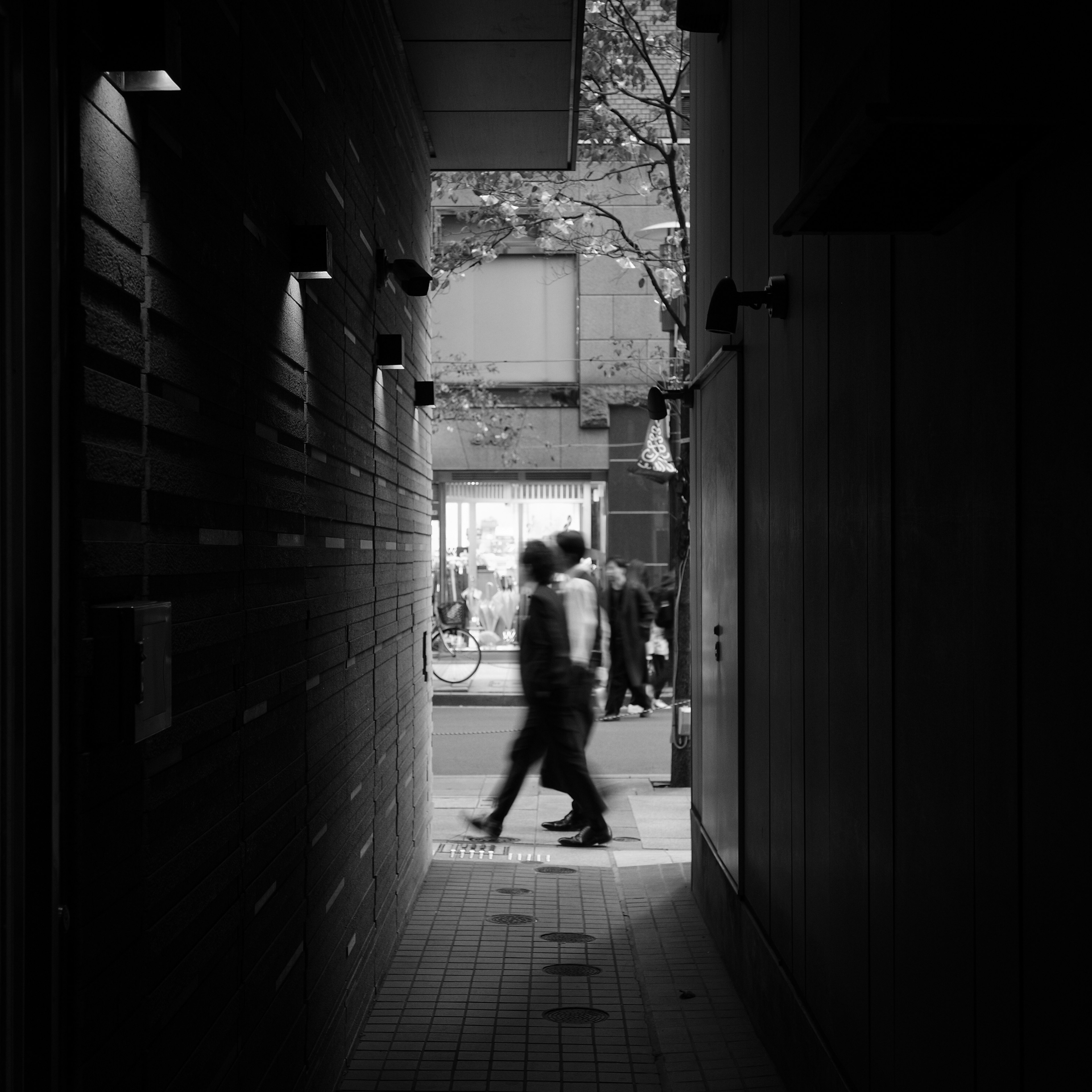 Silhouettes de personnes marchant dans un passage étroit avec une rue lumineuse en arrière-plan