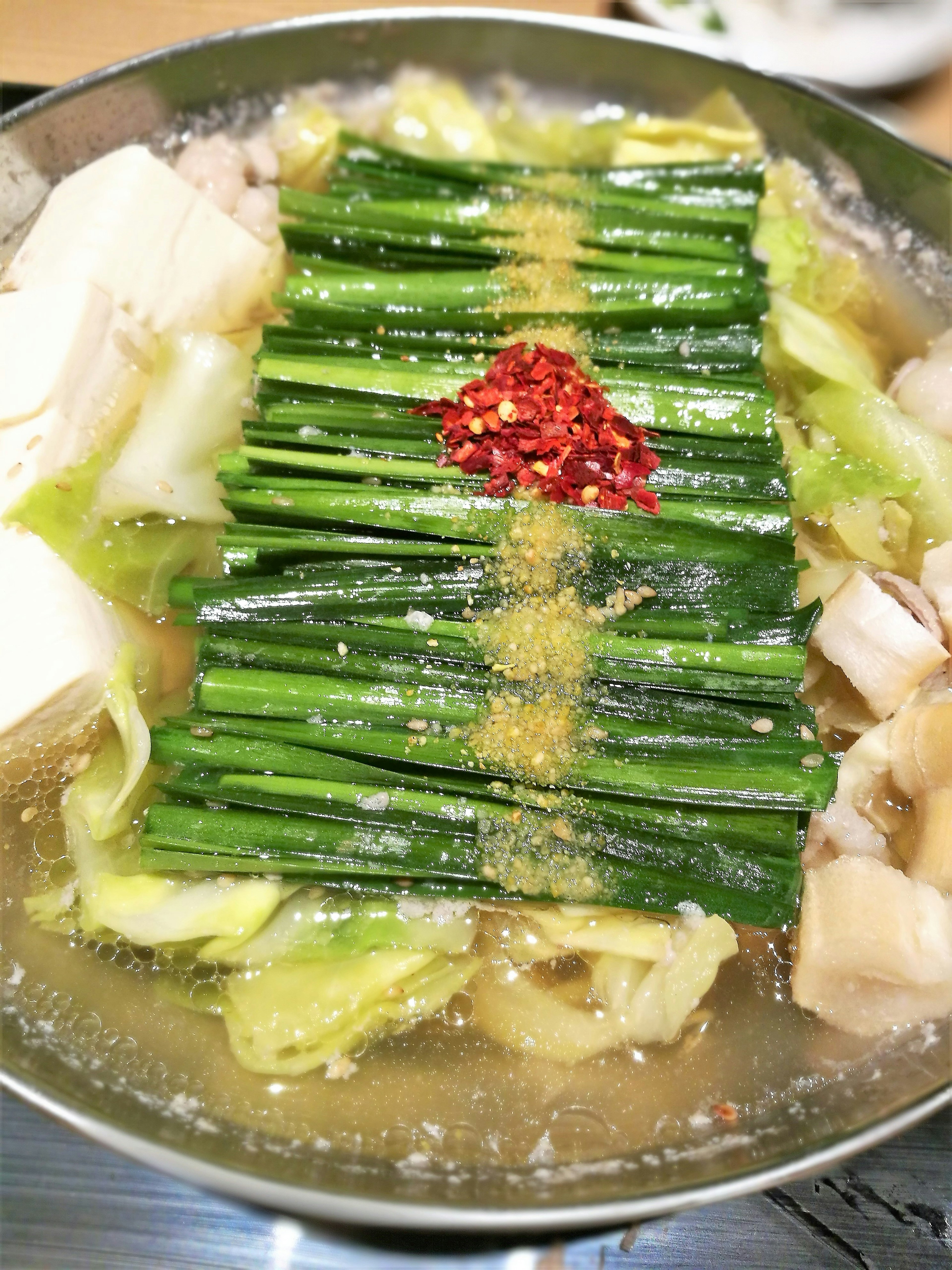 Hidangan hot pot yang menampilkan ayam, tahu, dan kol dengan daun bawang di atasnya dan hiasan cabai merah