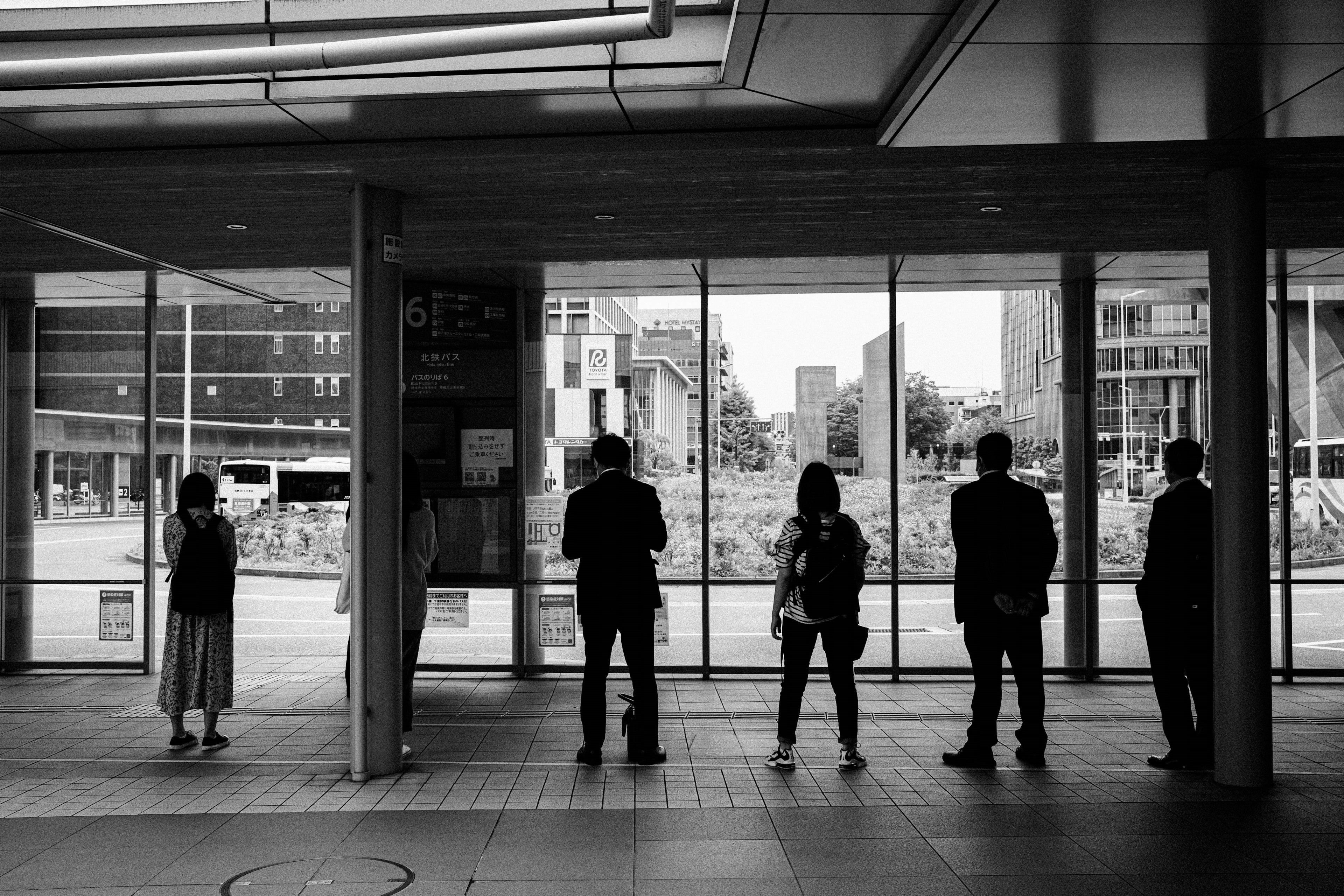 Personas de pie en una sala de espera en blanco y negro mirando por grandes ventanas