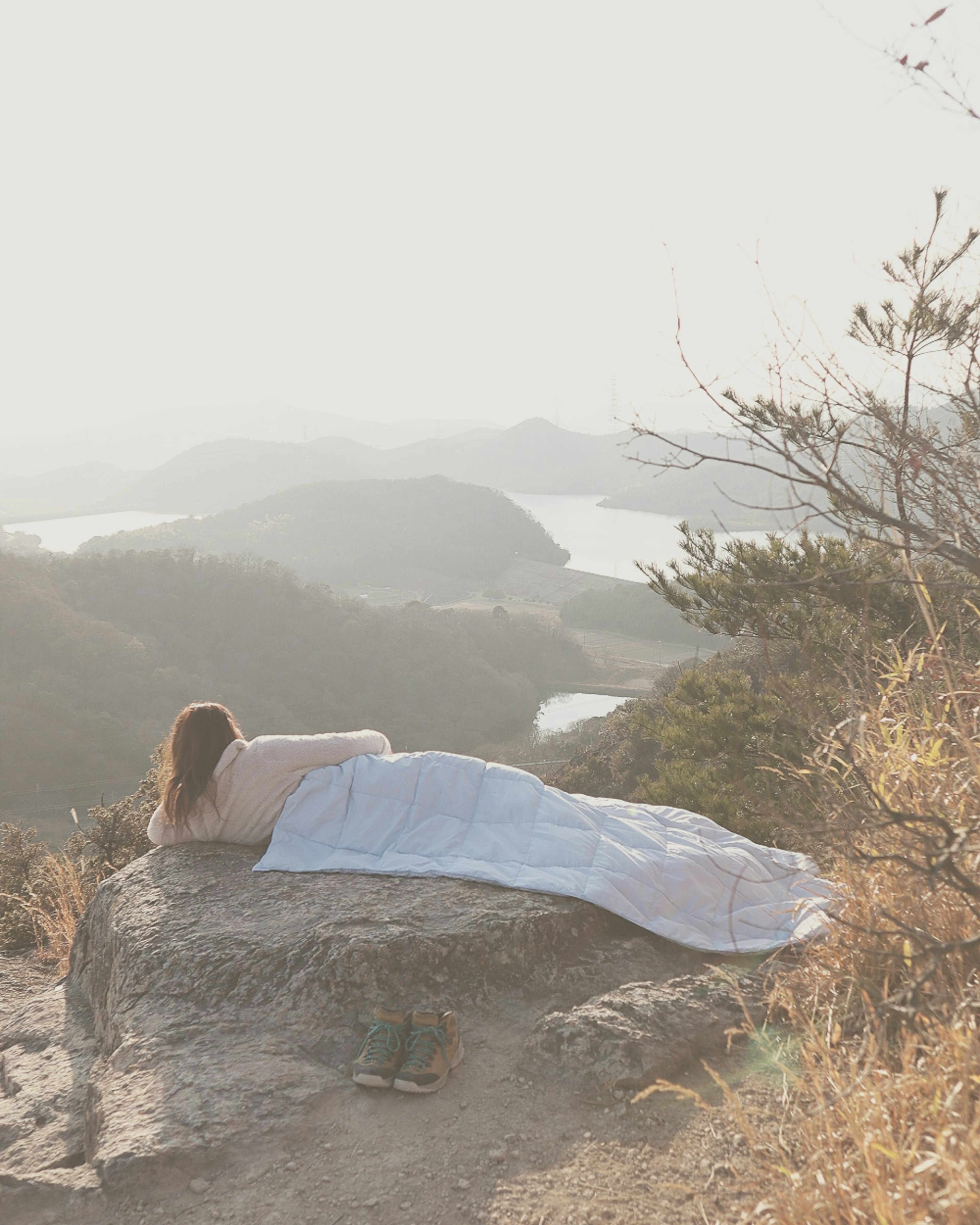 Woman lying on a rock covered with a blue blanket in a misty landscape overlooking water and hills