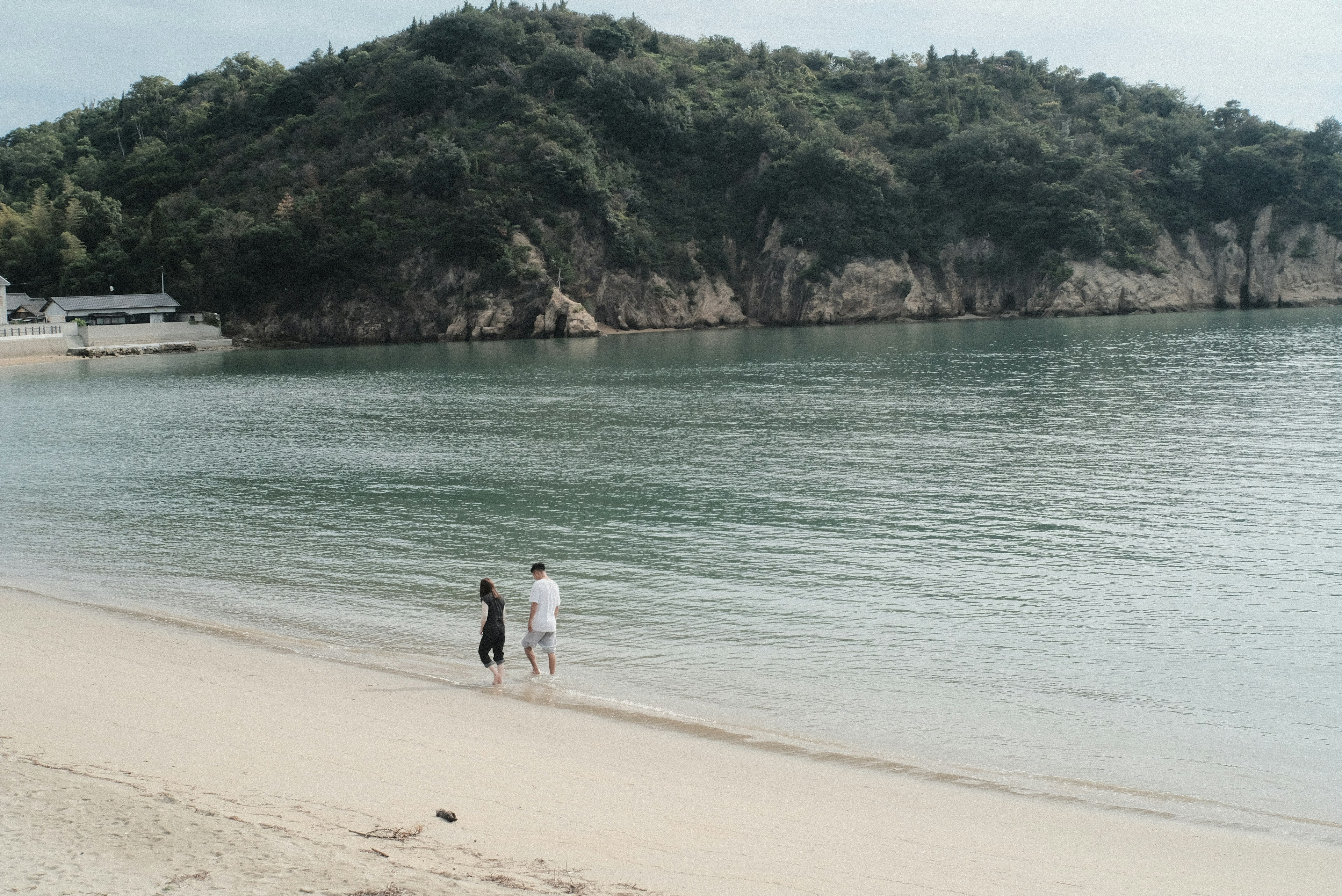 Dua orang berjalan di pantai dengan air tenang dan bukit di latar belakang