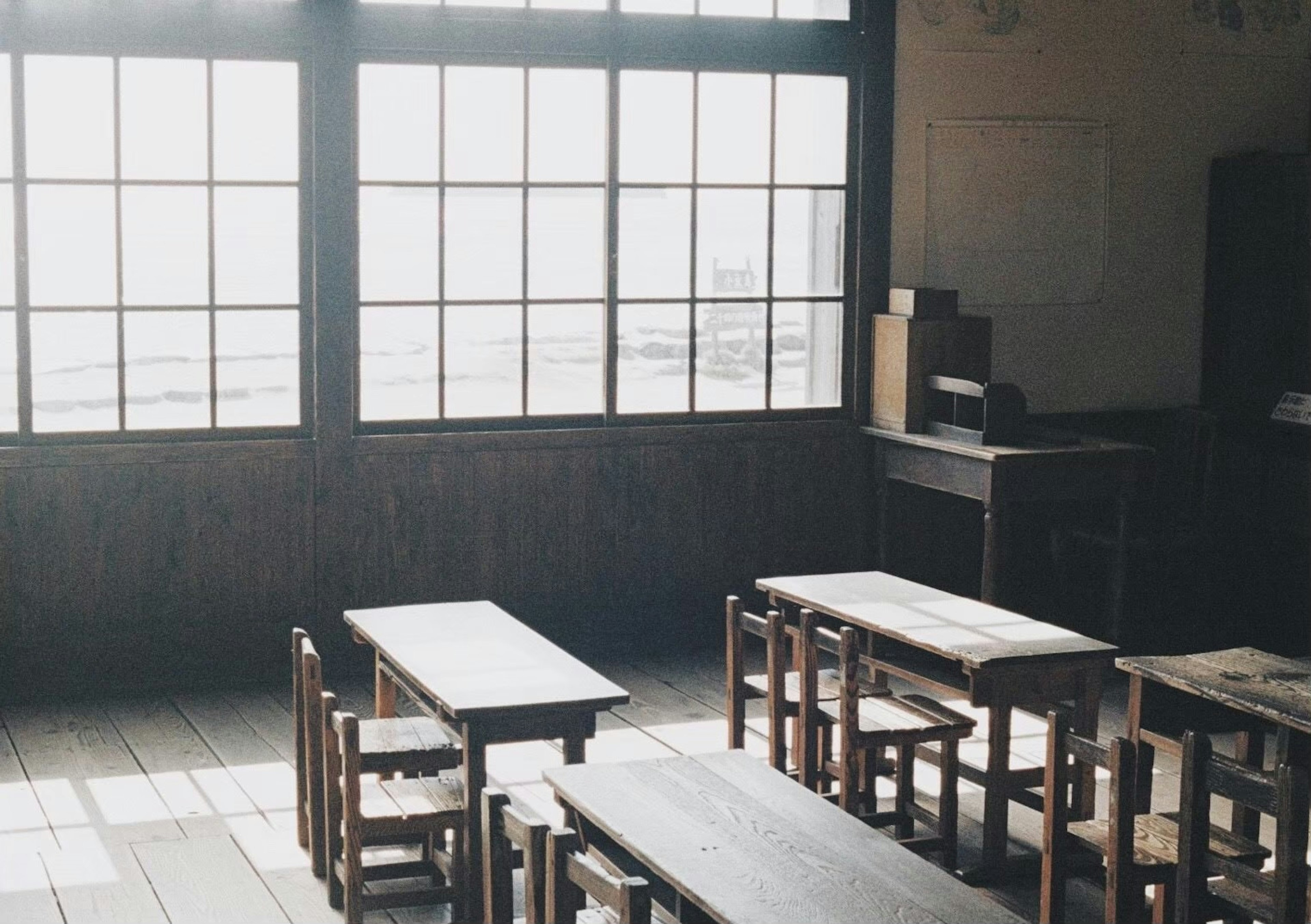 Innenansicht eines alten Klassenzimmers mit hellem Licht, das durch große Fenster strömt, Holztische und Stühle ordentlich angeordnet