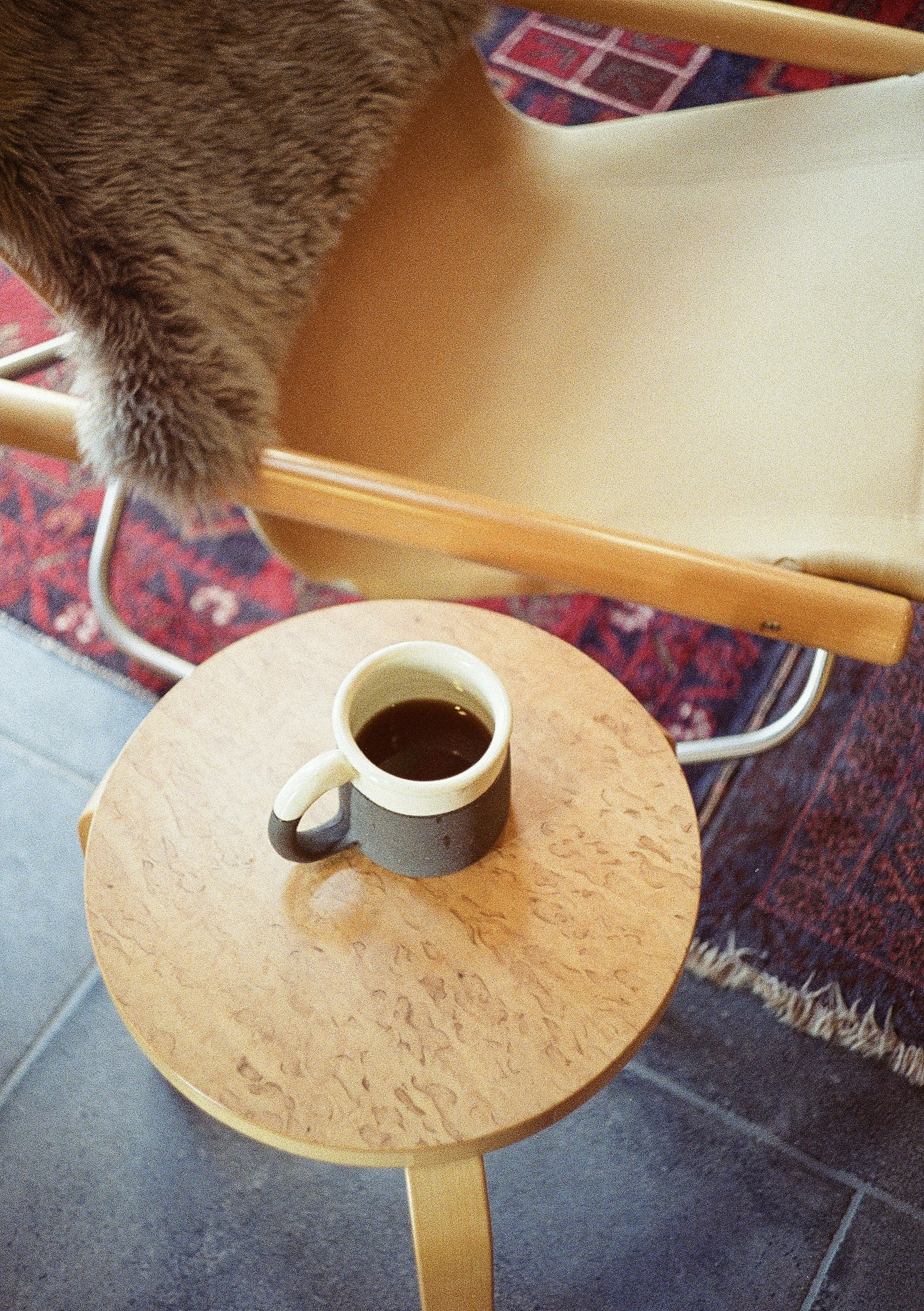 Una taza de café sobre una mesa de madera con parte de una silla y una manta en un entorno interior simple