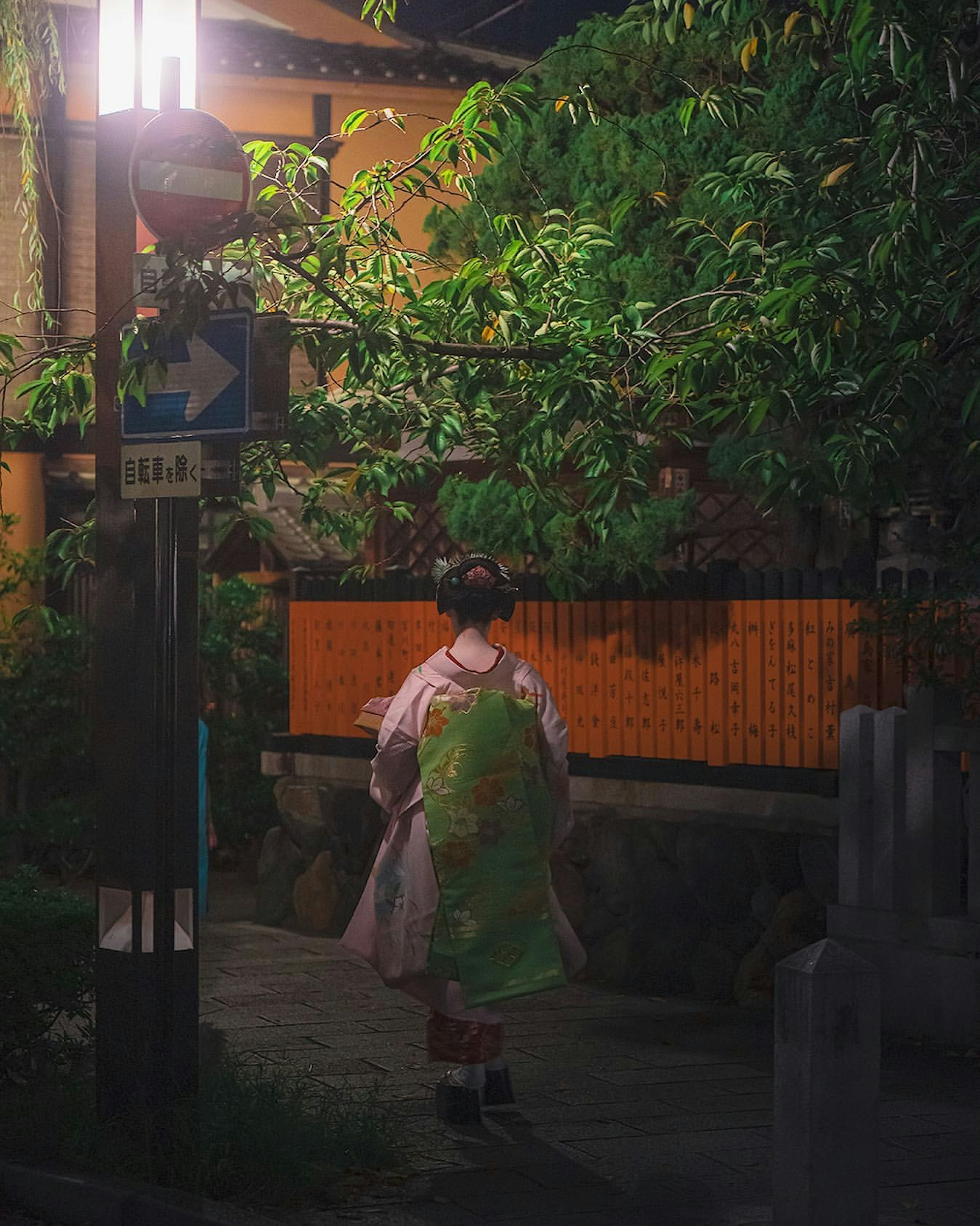 Femme en kimono marchant dans la rue la nuit avec un lampadaire et une végétation luxuriante