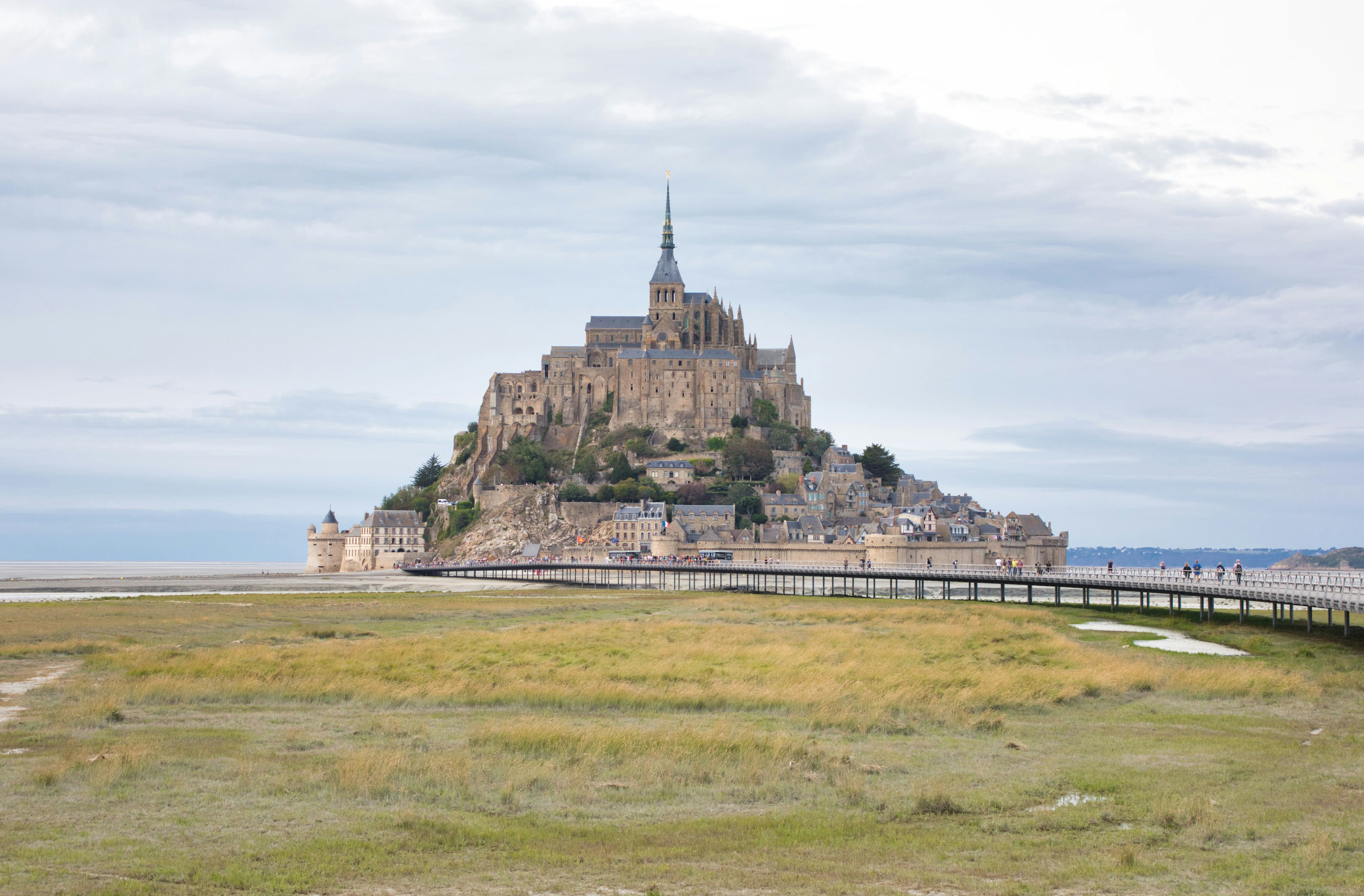 Pemandangan Mont Saint-Michel dengan lanskap berumput dan jembatan di bawah langit mendung