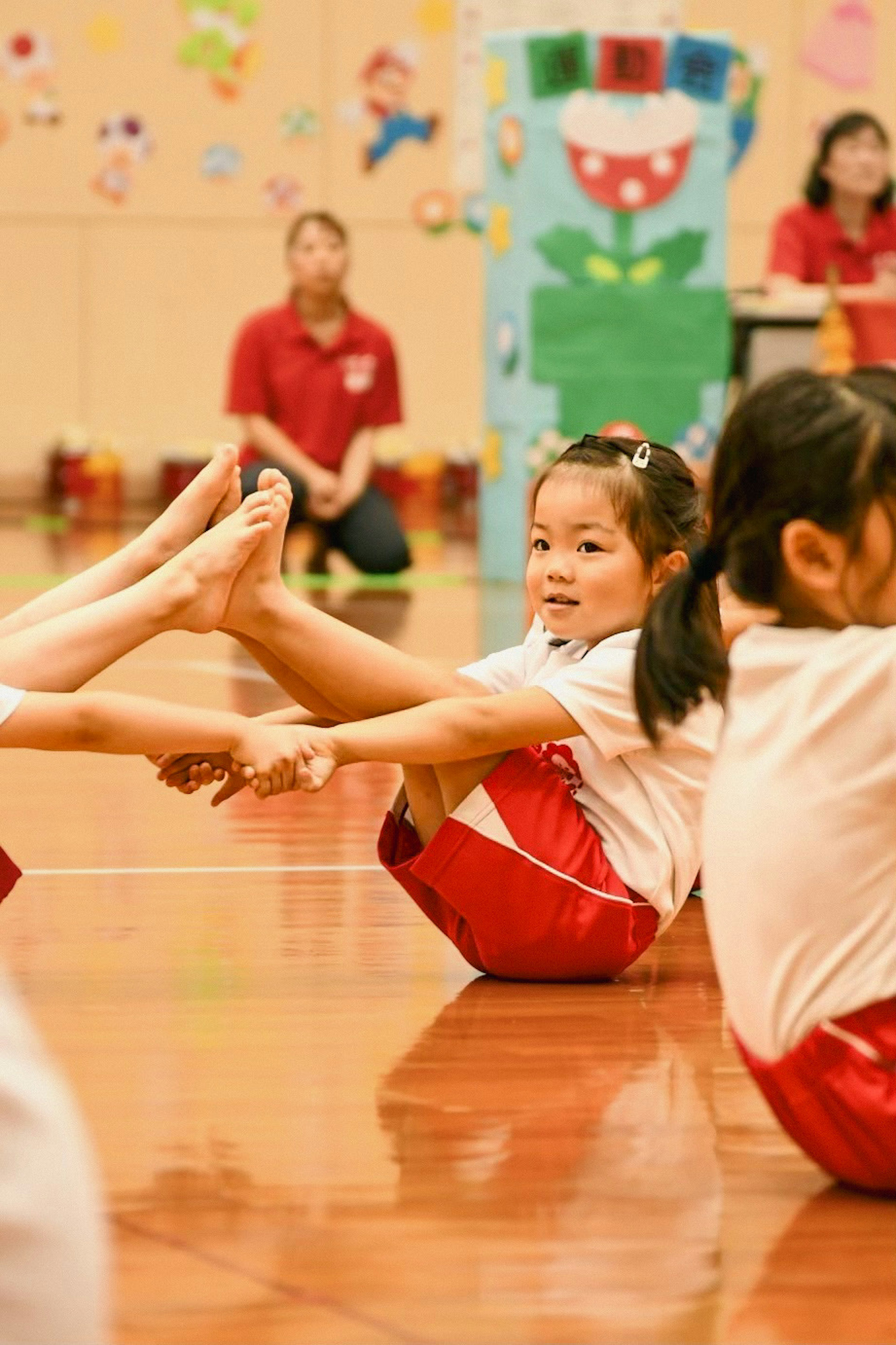 子供たちが体育館で体操をしている様子 赤いユニフォームを着た女の子がポーズをとっている 背景にはカラフルな装飾がある