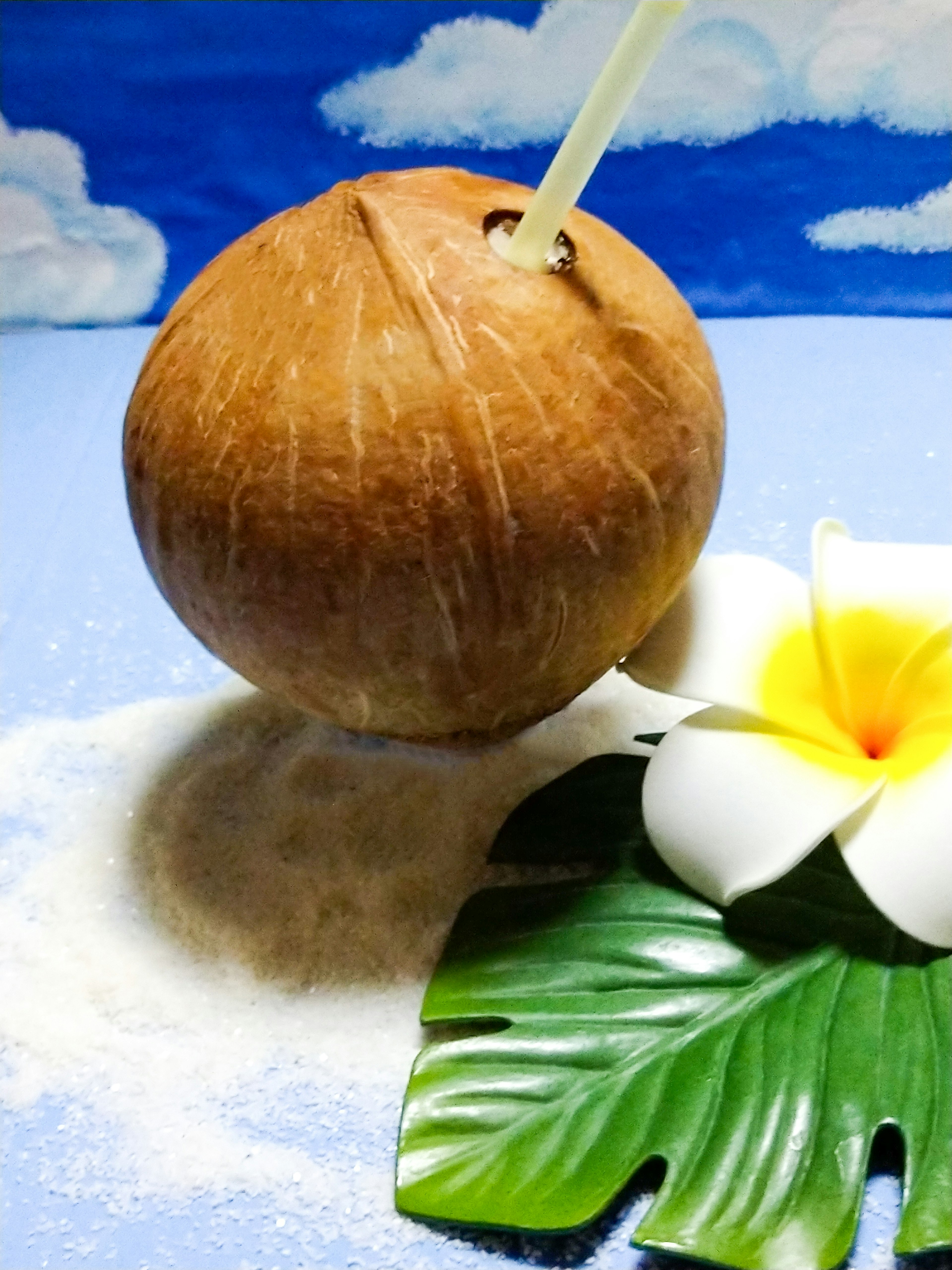 Cocco con una cannuccia accanto a un fiore di plumeria su uno sfondo di spiaggia sabbiosa