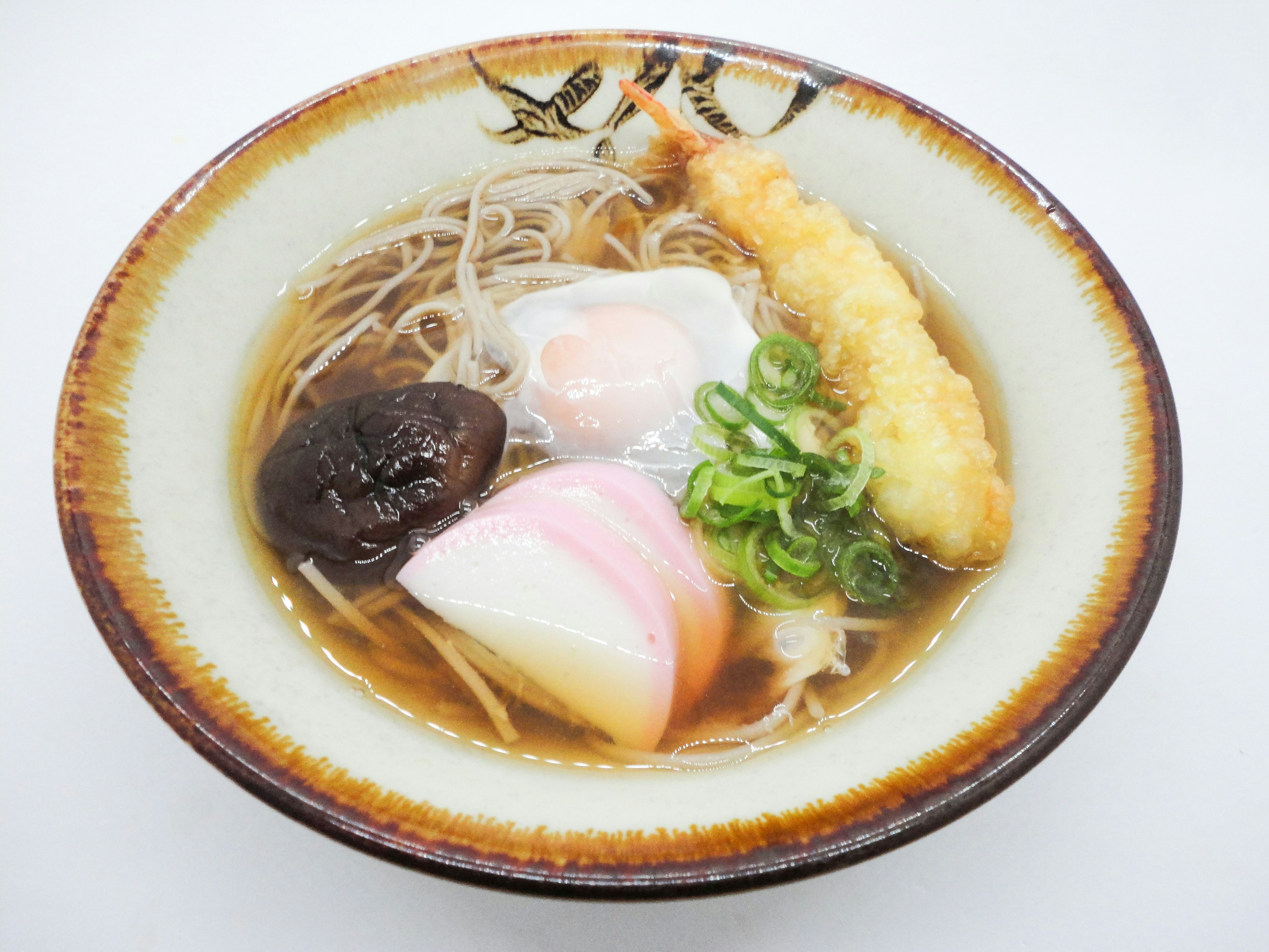 A bowl of warm soba soup topped with shrimp tempura, green onions, kamaboko, and shiitake mushrooms