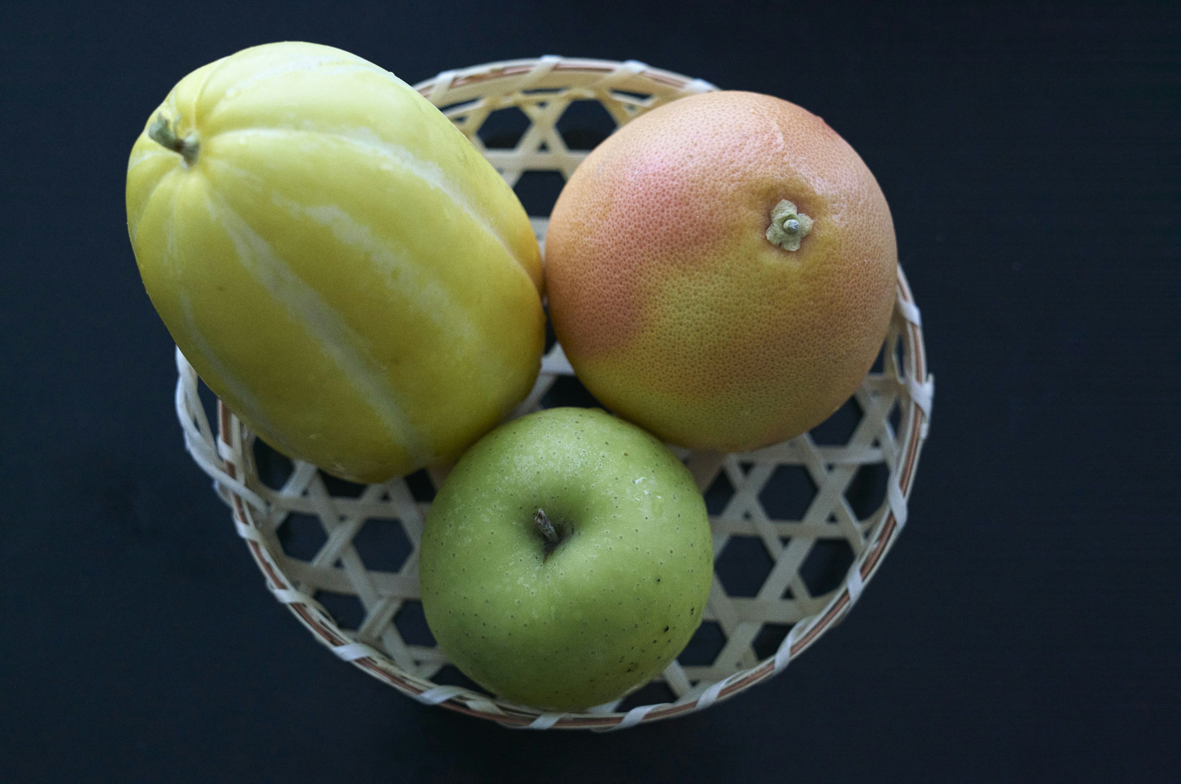 A wicker basket containing a yellow melon an orange fruit and a green apple