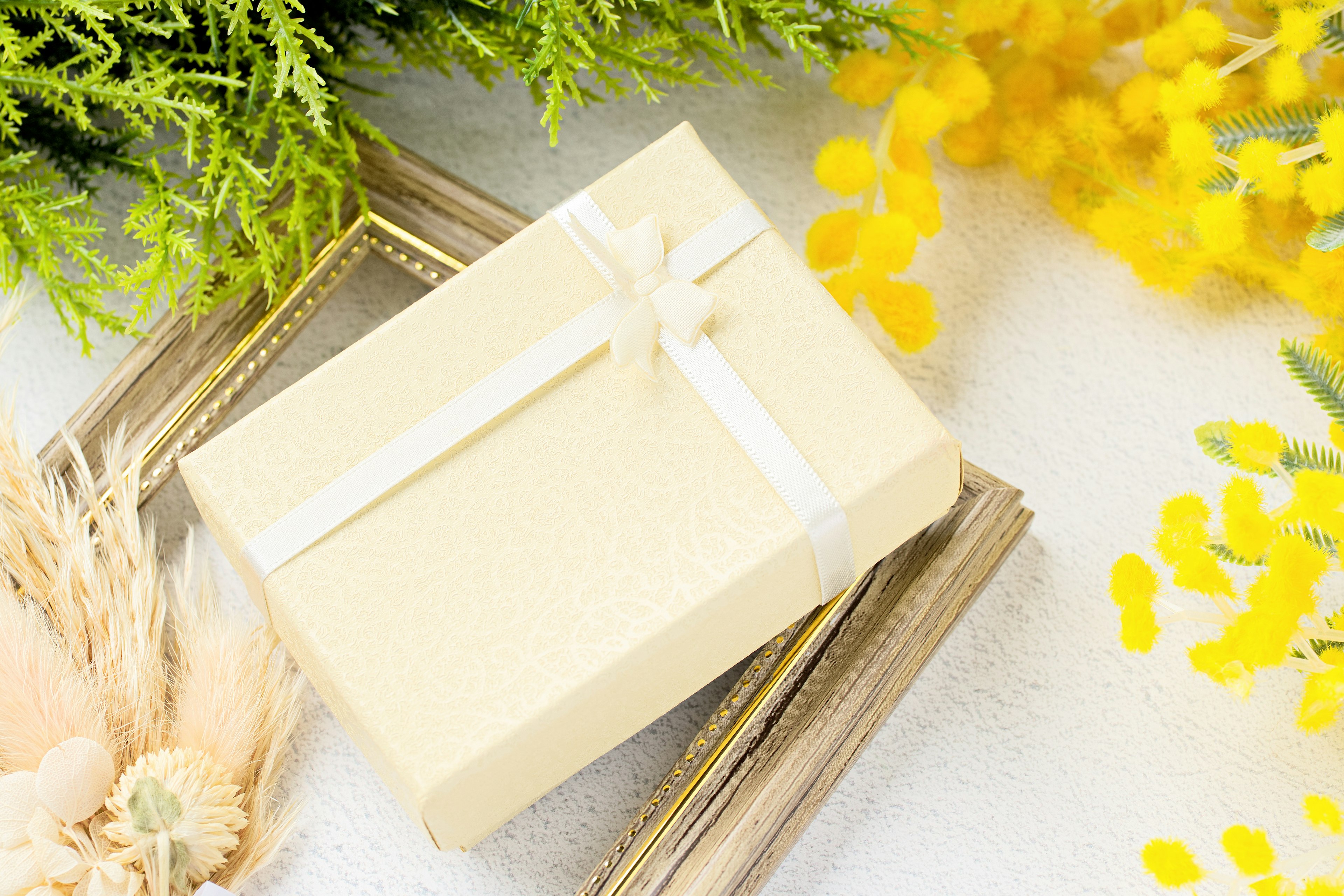 Caja de regalo color crema con una cinta blanca rodeada de flores amarillas y follaje verde