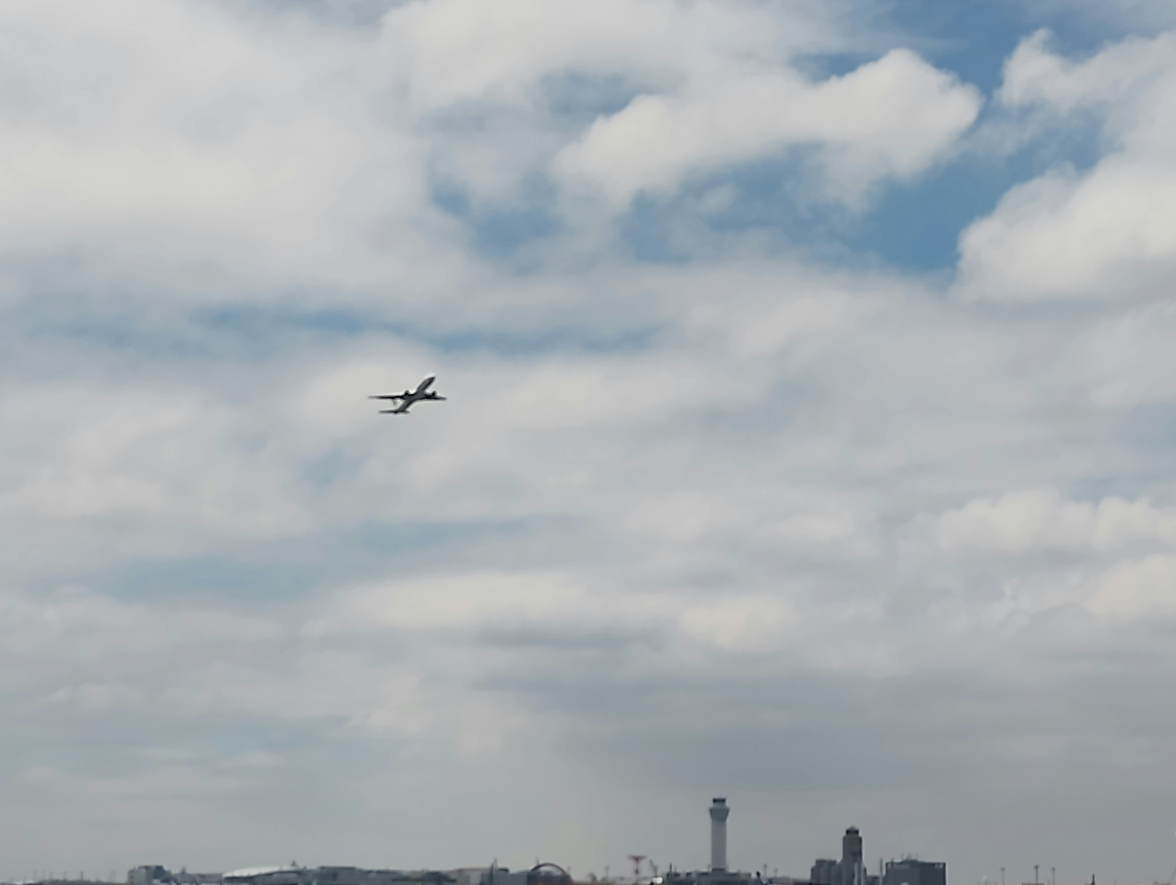 Un avión volando en un cielo azul con nubes y una torre de aeropuerto visible a lo lejos