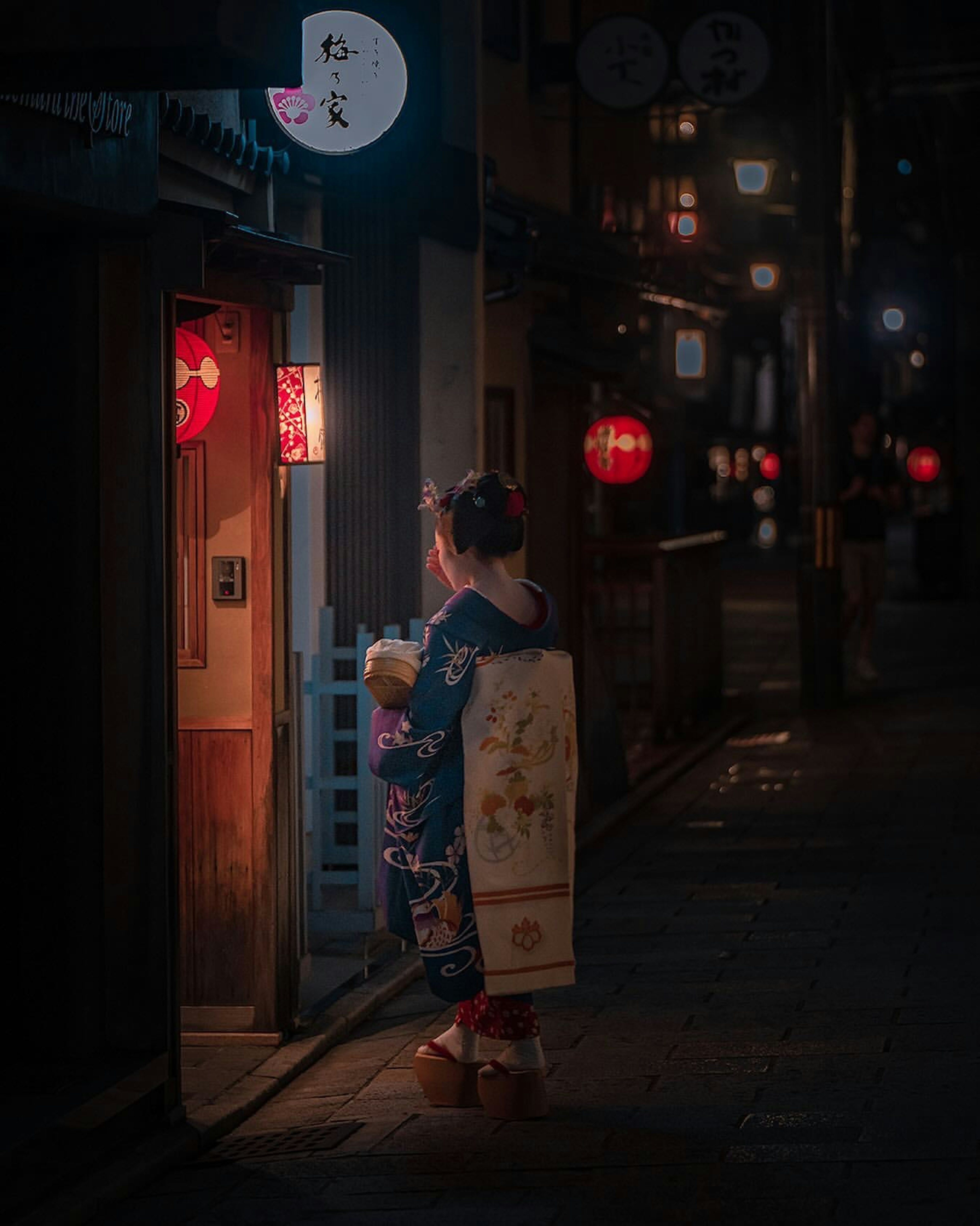 Eine Frau in einem Kimono steht vor einem hell erleuchteten Geschäft in einer nächtlichen Straße