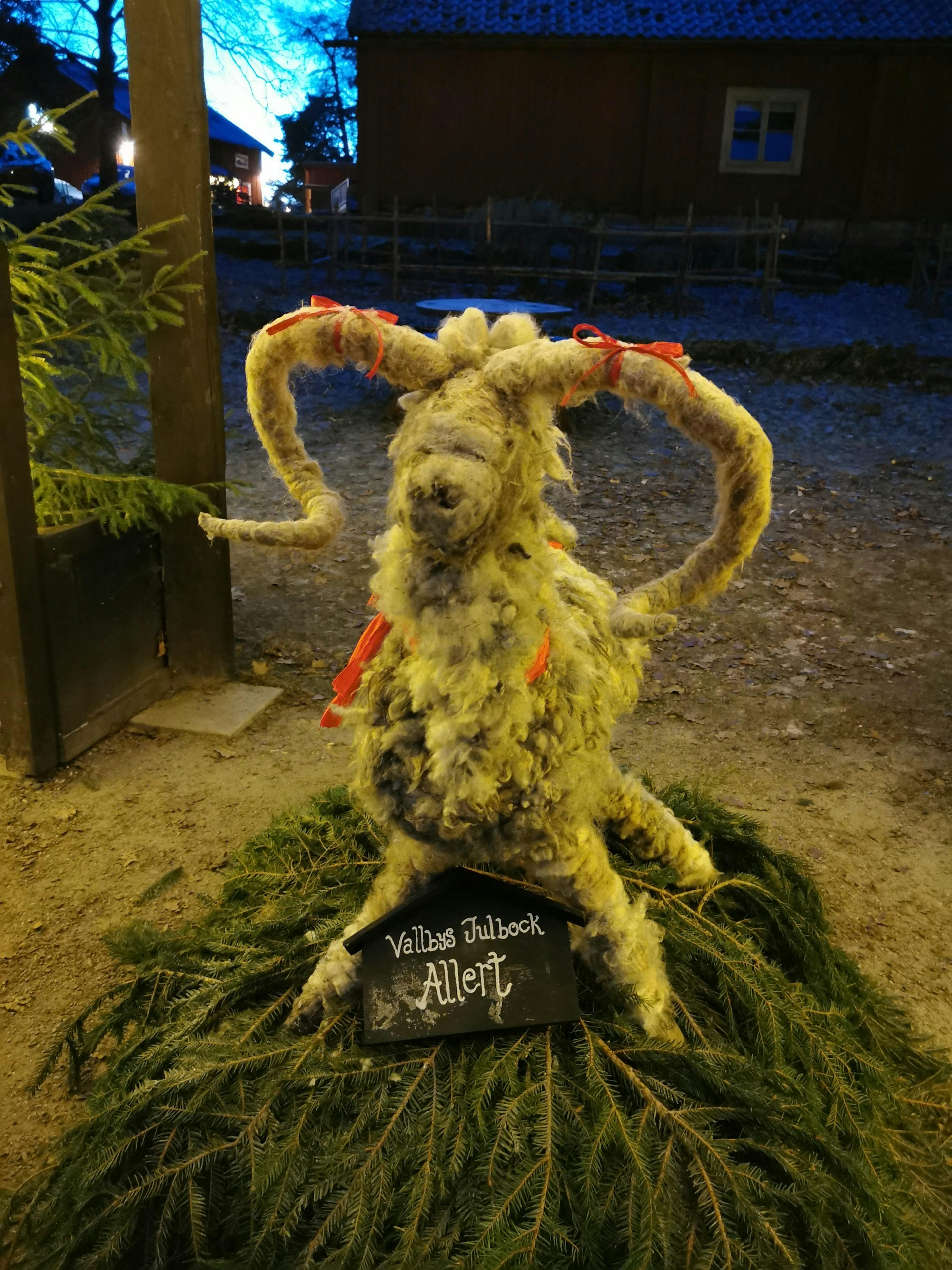 Estatua de cabra hecha de paja con cuernos enrollados colocada sobre vegetación en un entorno nocturno