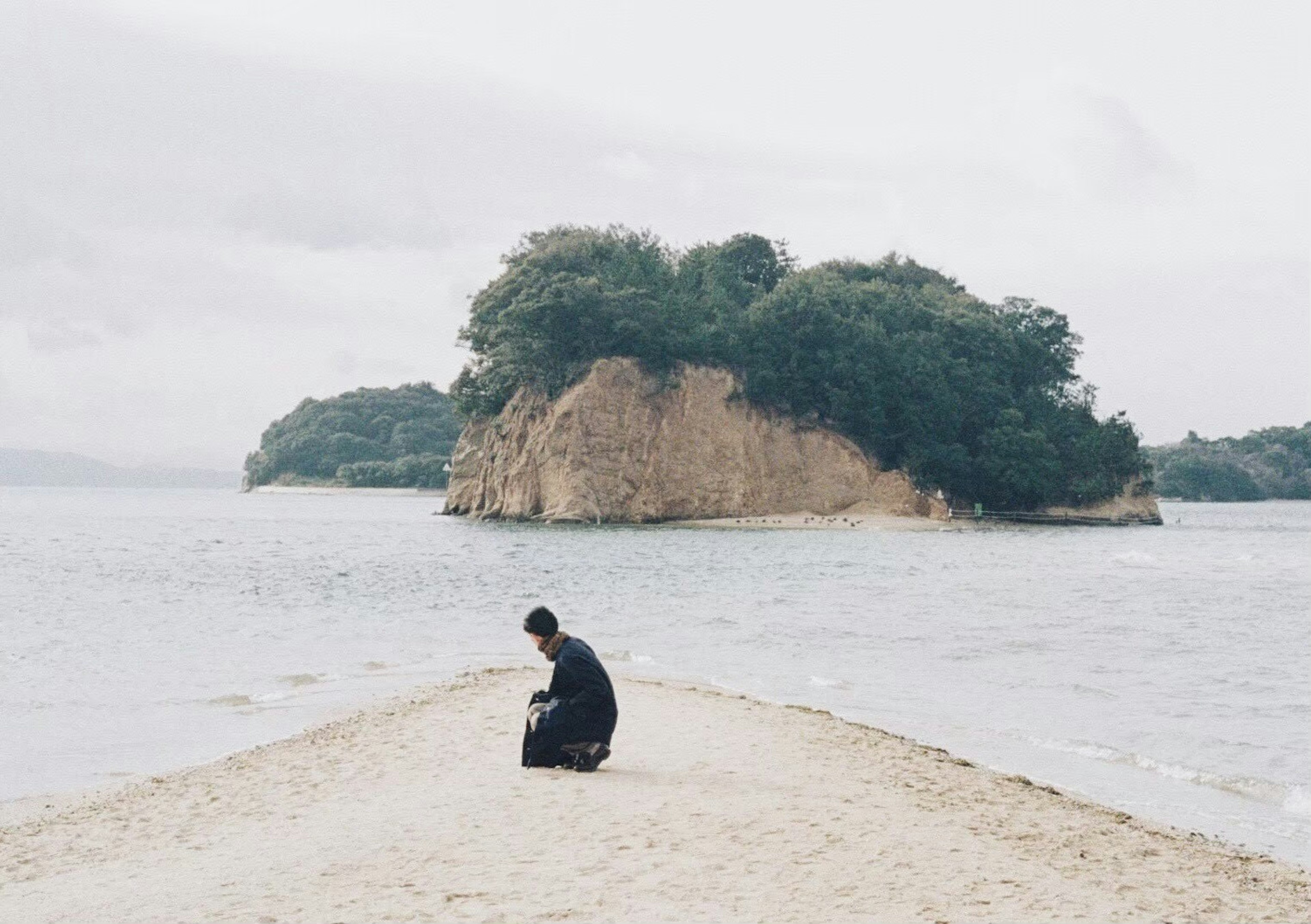 Person in black clothing sitting on a beach with a small island in the background