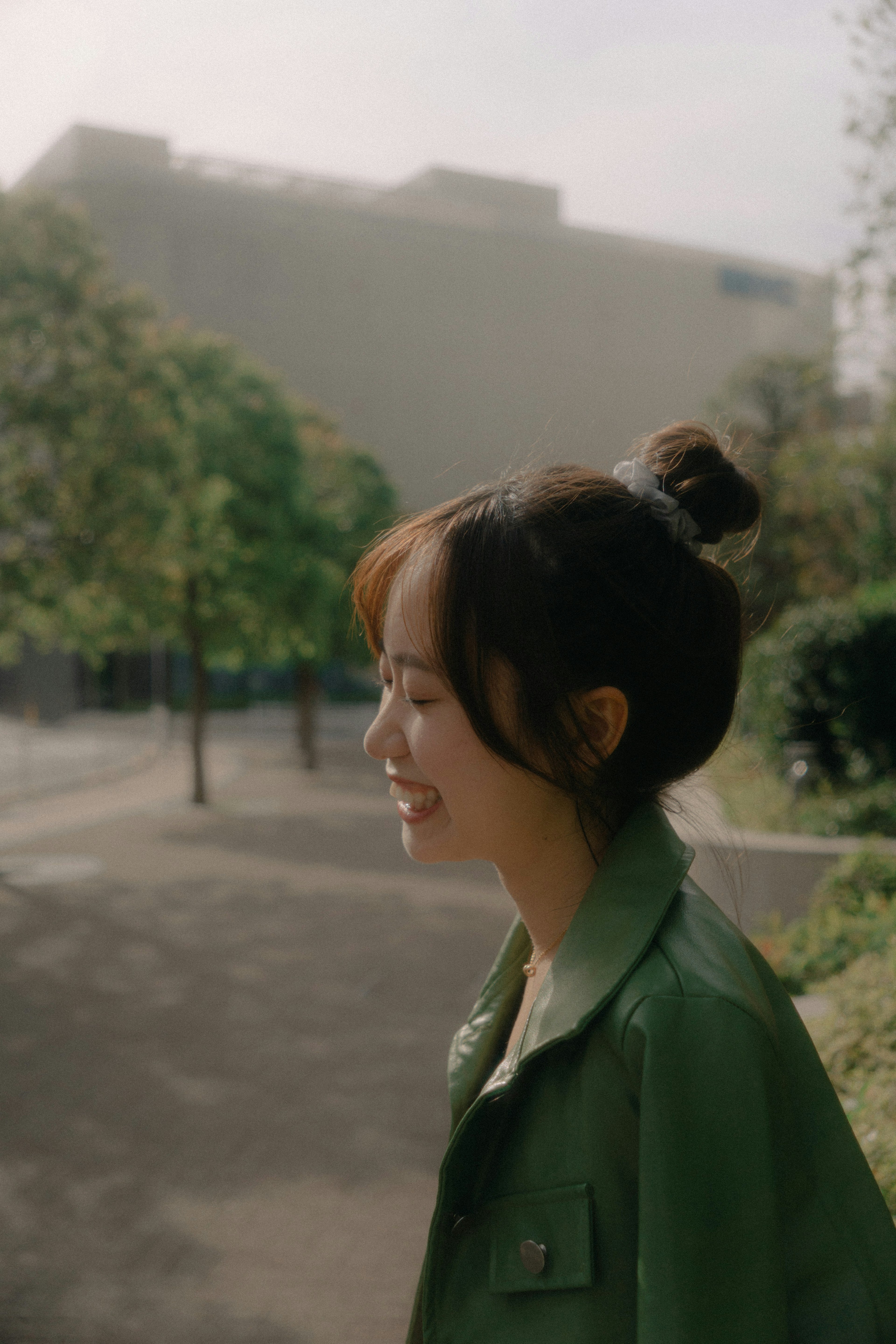 Profile of a smiling woman in a green jacket standing in a park with trees and a building in the background