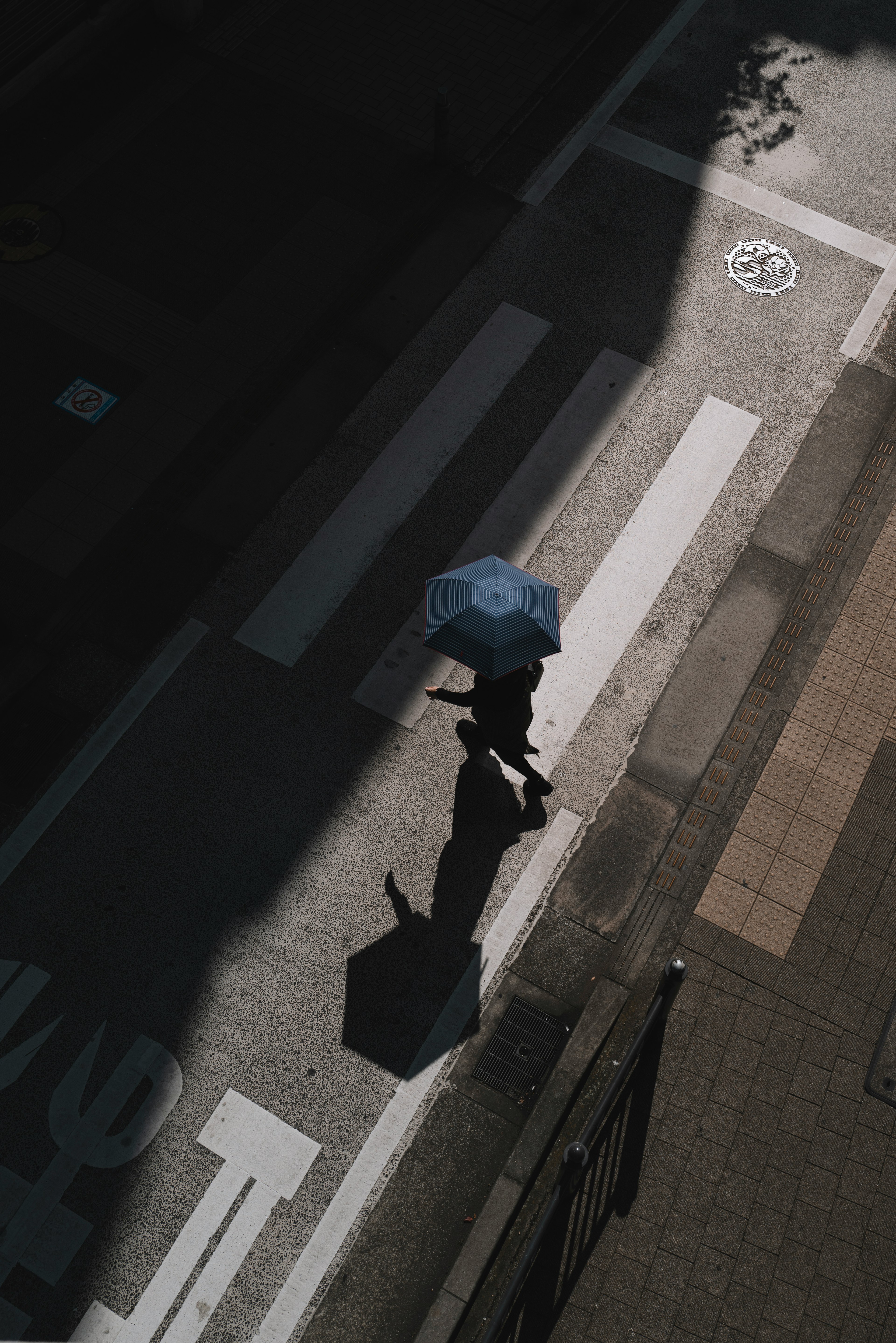 A person with an umbrella walking across a crosswalk in shadow