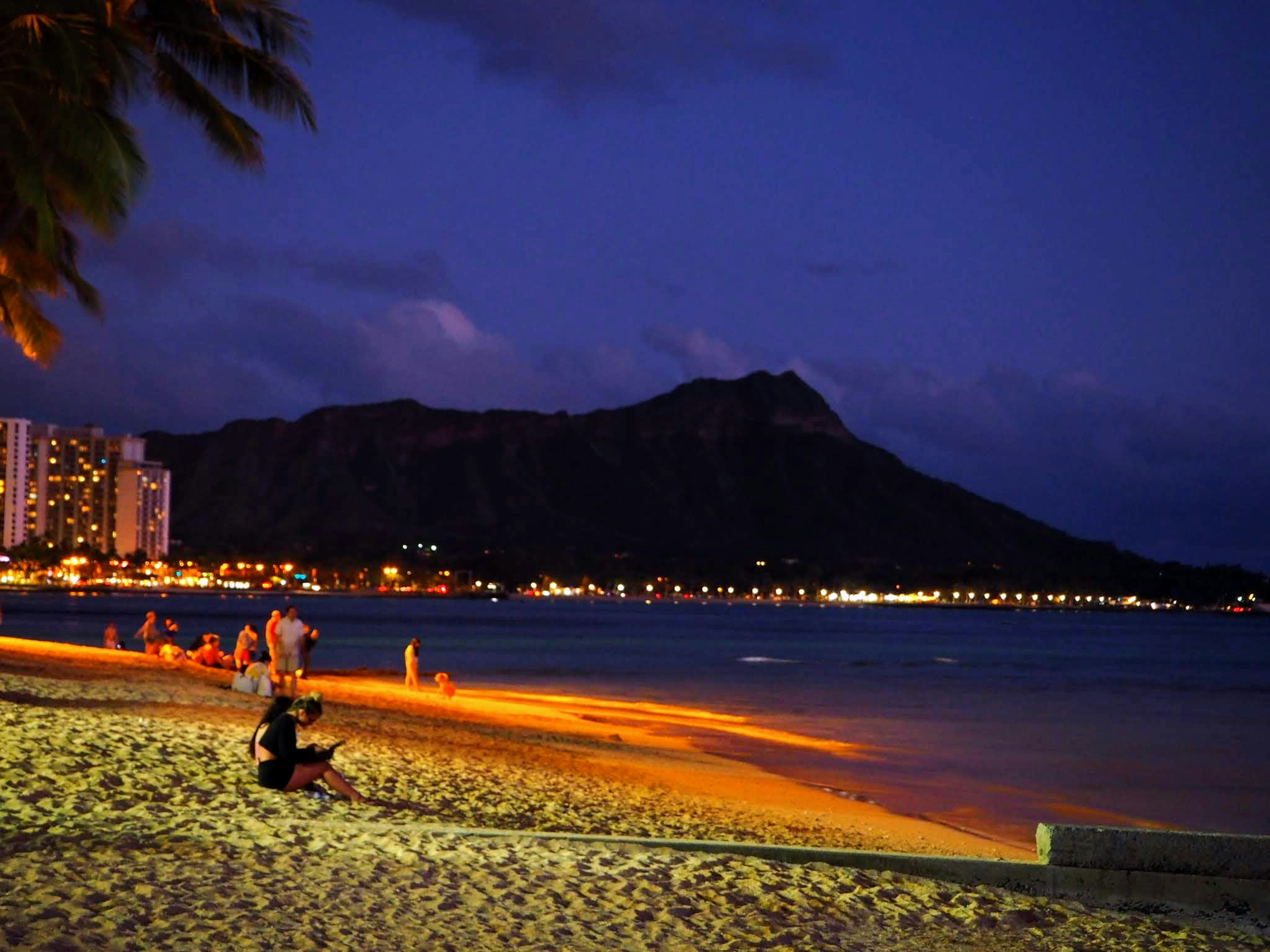 Pemandangan pantai malam dengan orang-orang duduk di pasir Diamond Head di latar belakang