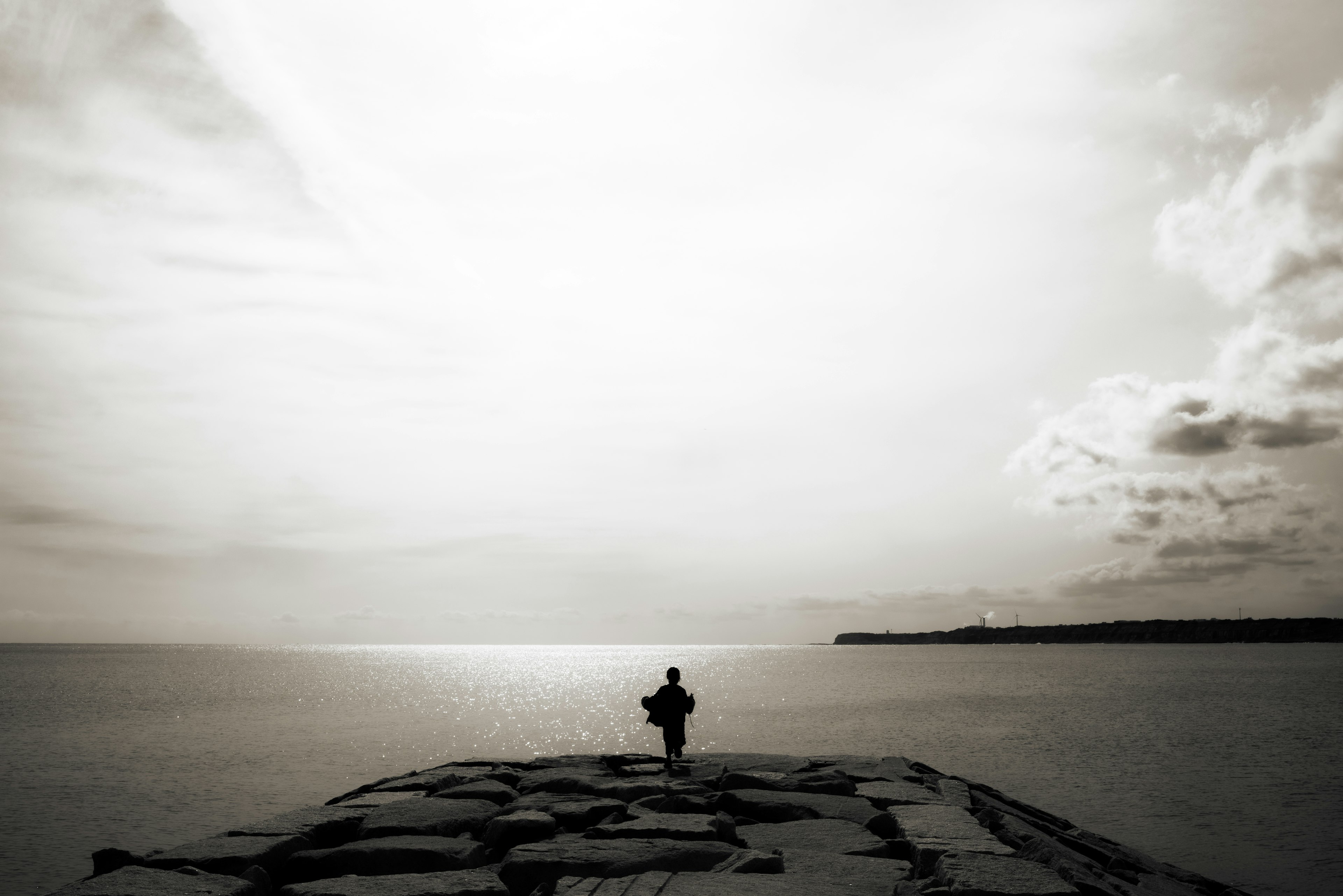 Schwarzweißbild einer Person, die auf einem Pier steht und auf das Meer unter einem weiten Himmel blickt