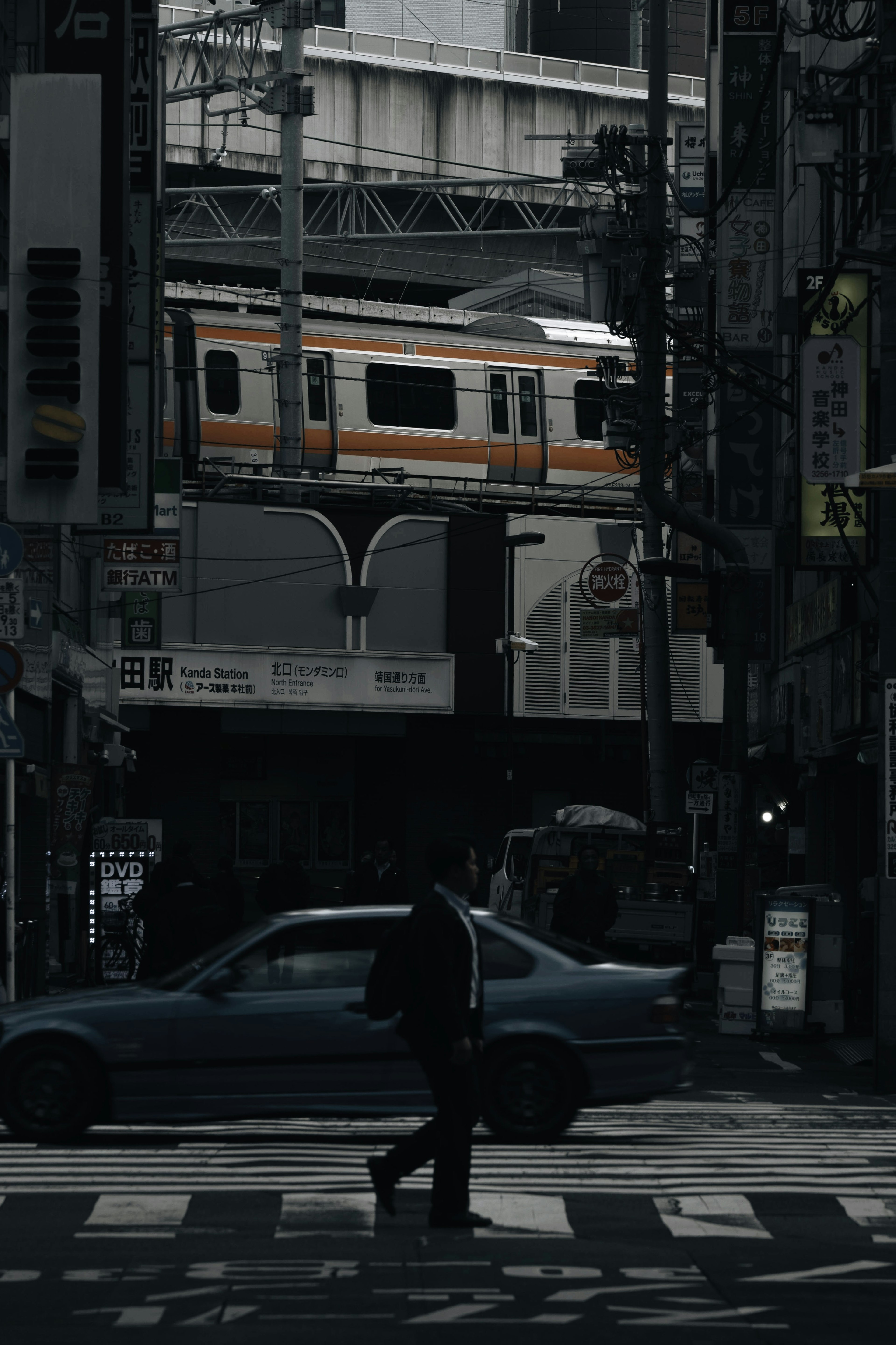 Dimly lit urban scene with an orange train passing overhead and a person crossing the street