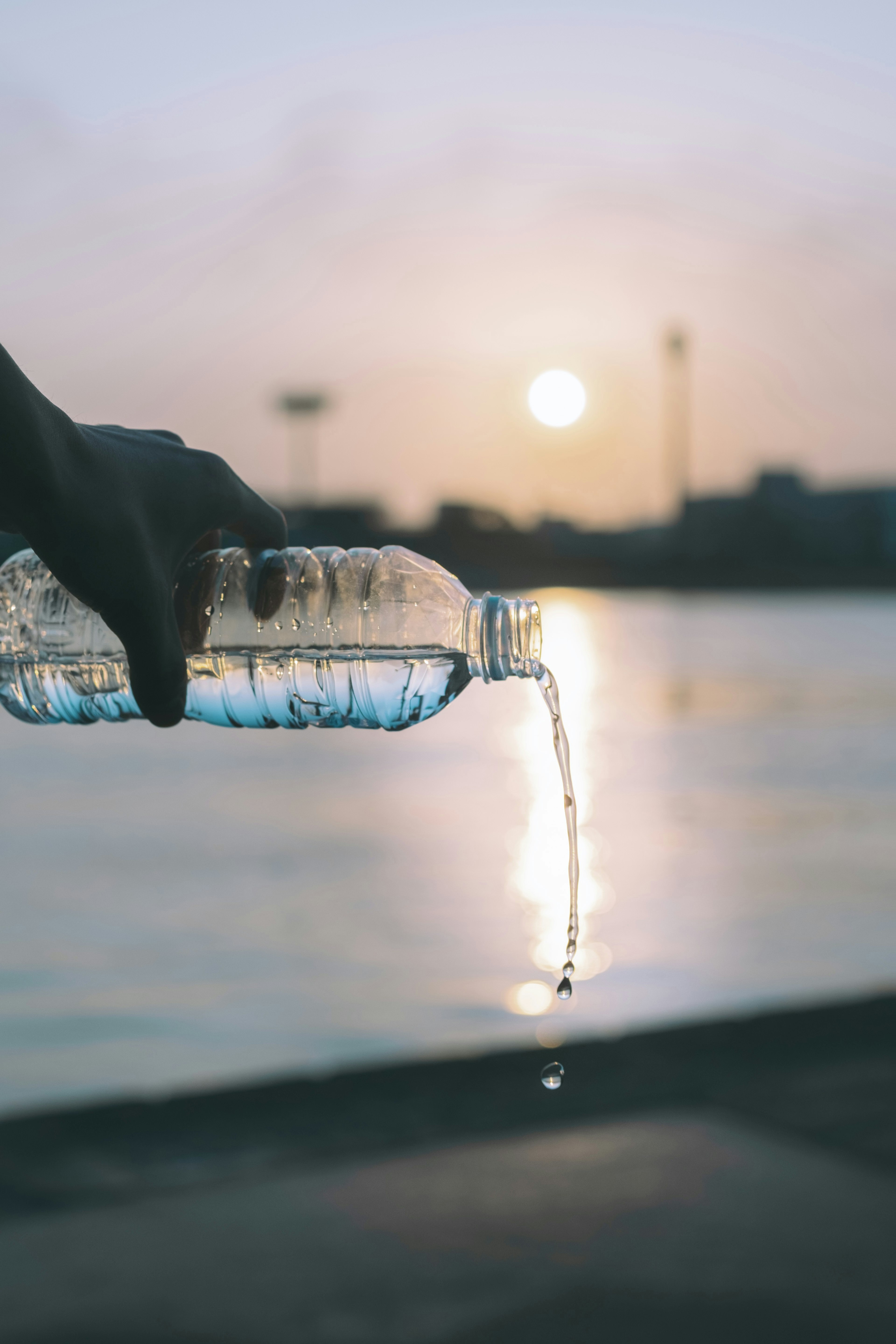 Una mano vertiendo agua de una botella contra un atardecer sobre una superficie de agua tranquila