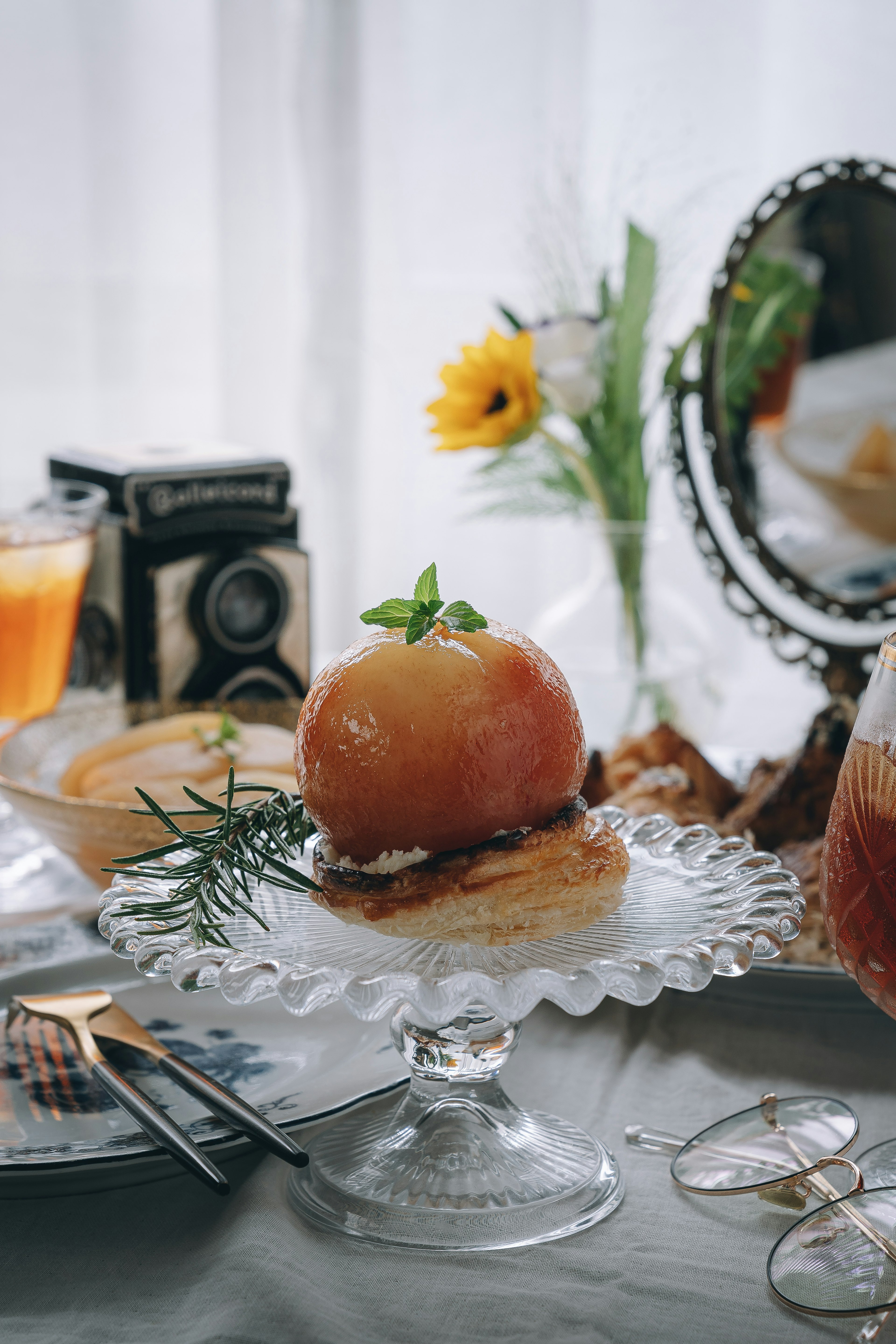 Un postre de manzana bellamente presentado en un soporte de vidrio con flores y una cámara vintage de fondo