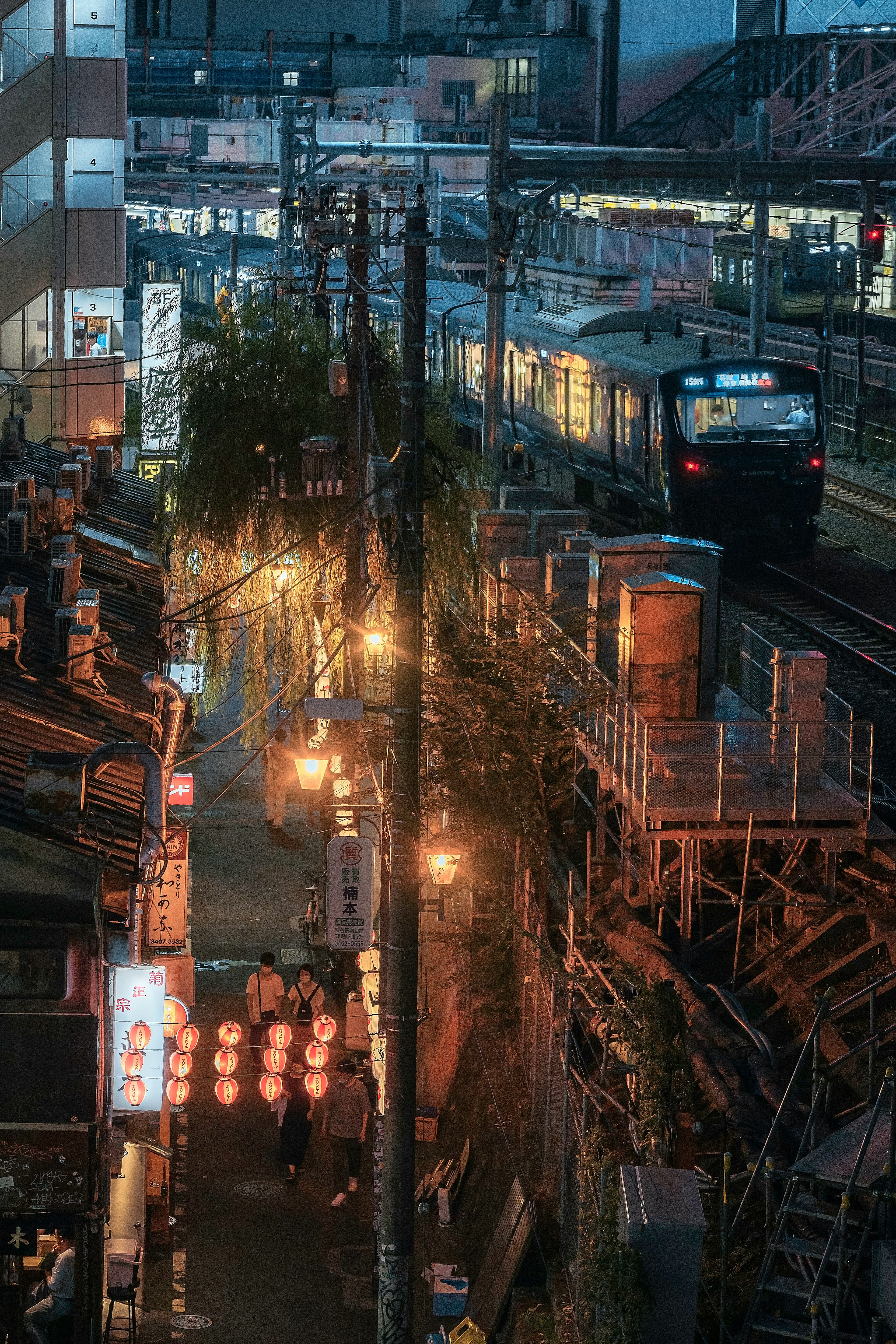 夜の日本の街並み、線路沿いの小道と提灯が並ぶ飲食店を背景に、列車が通過する風景