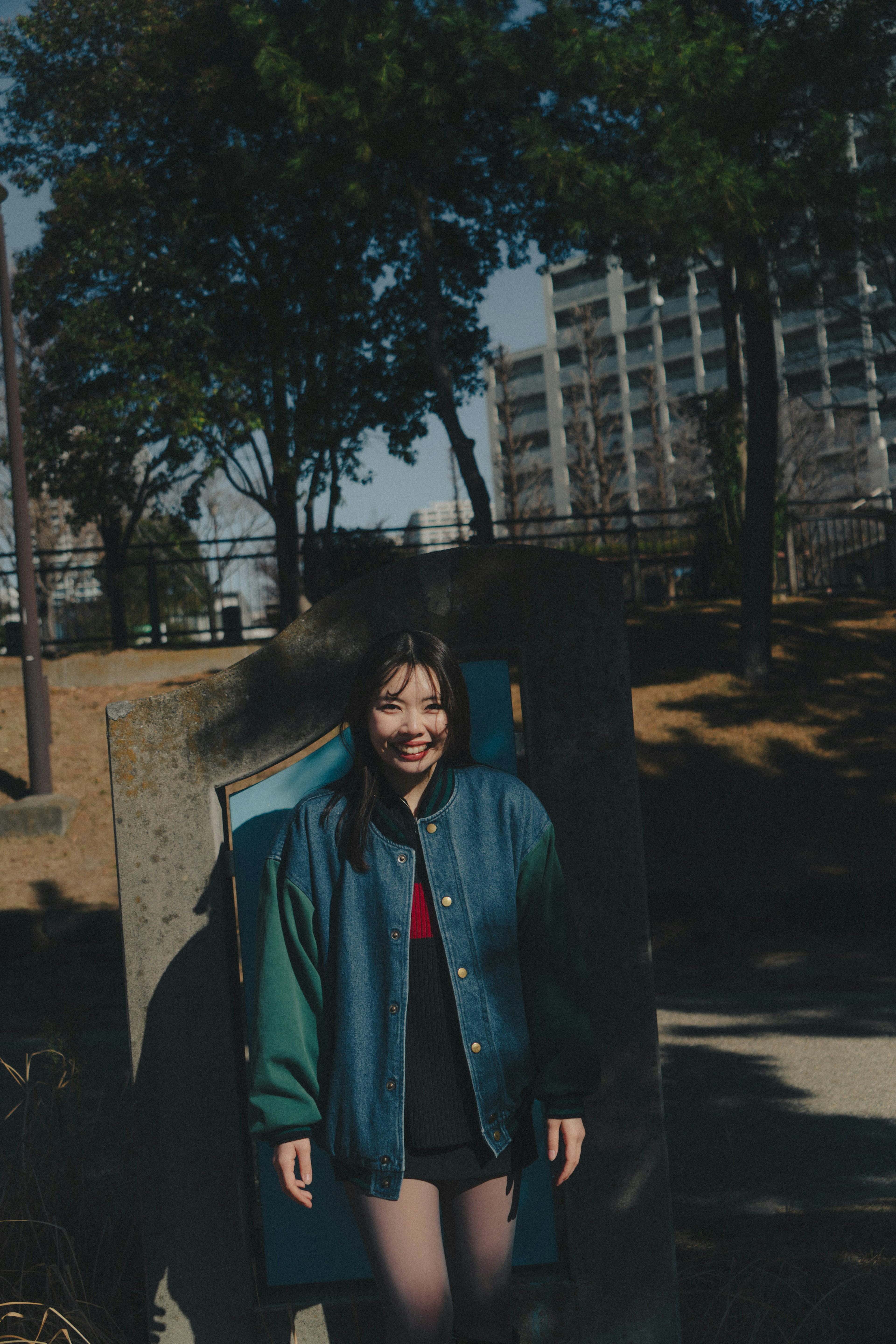 Woman smiling in a park wearing a teal jacket with buildings and trees in the background