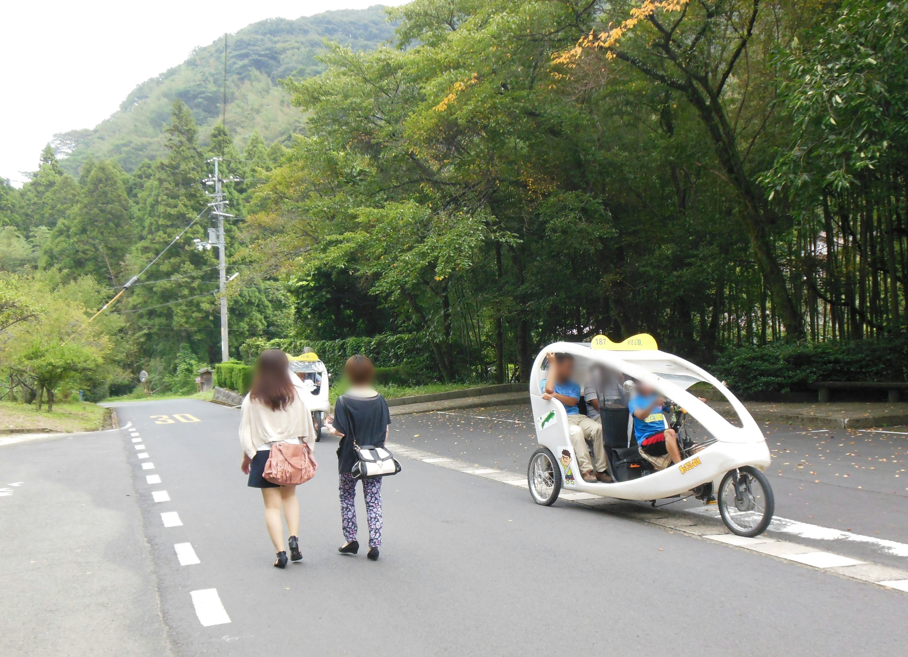 Two women walking on a tree-lined road with a white bicycle taxi