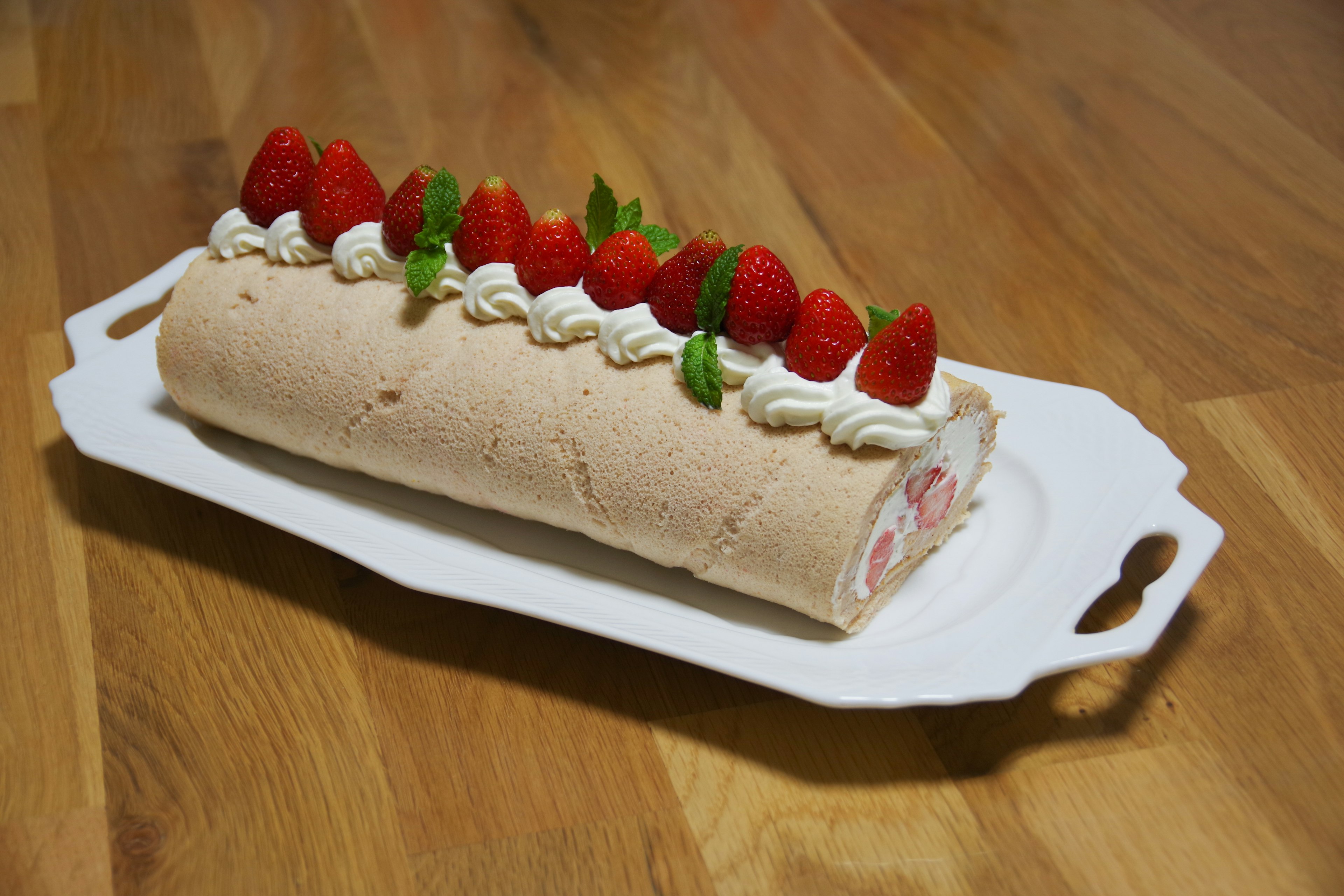 A rolled cake topped with strawberries and whipped cream served on a white platter