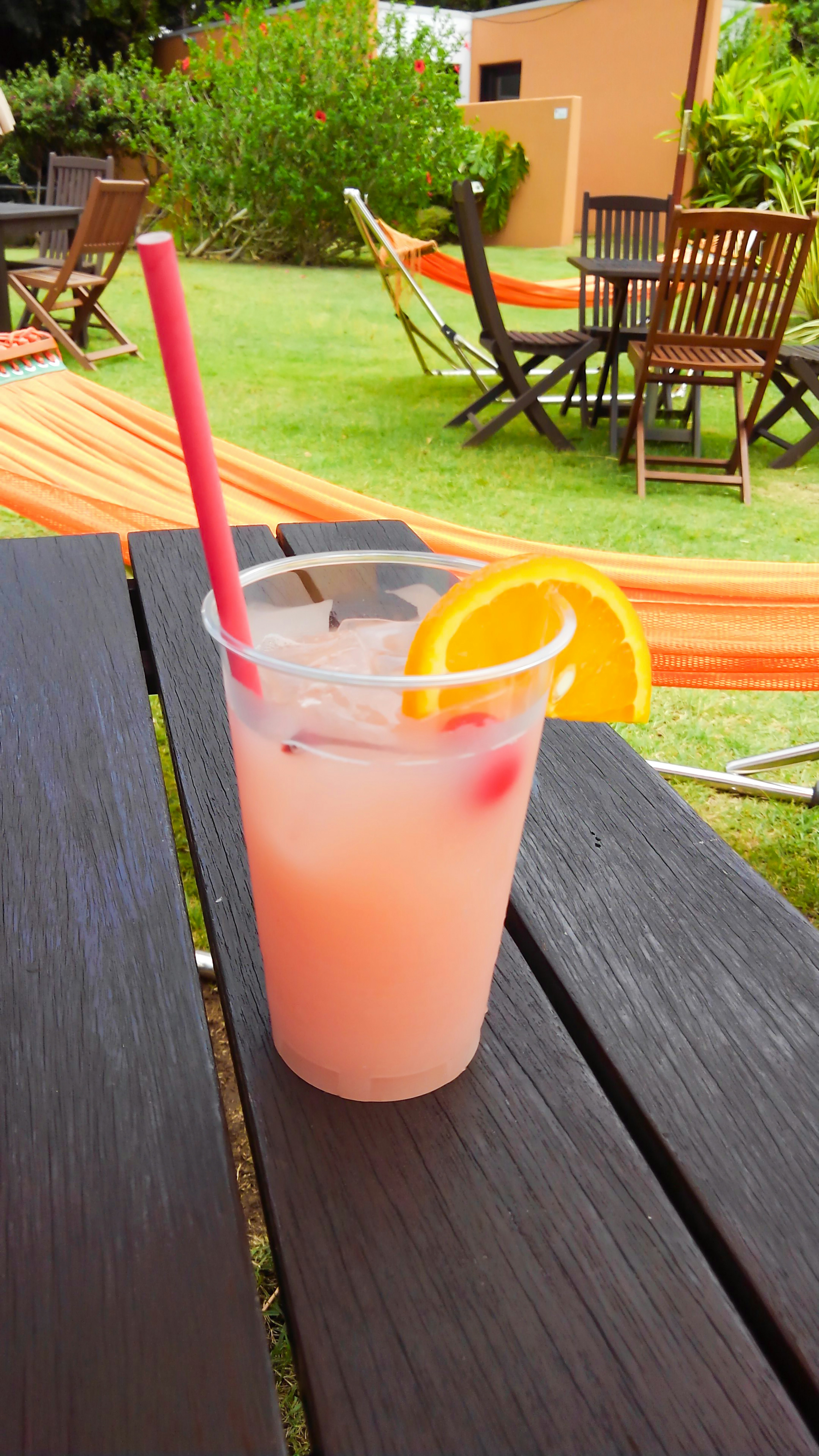 A cocktail glass with an orange slice and a red straw on a wooden table with hammocks and chairs in the background