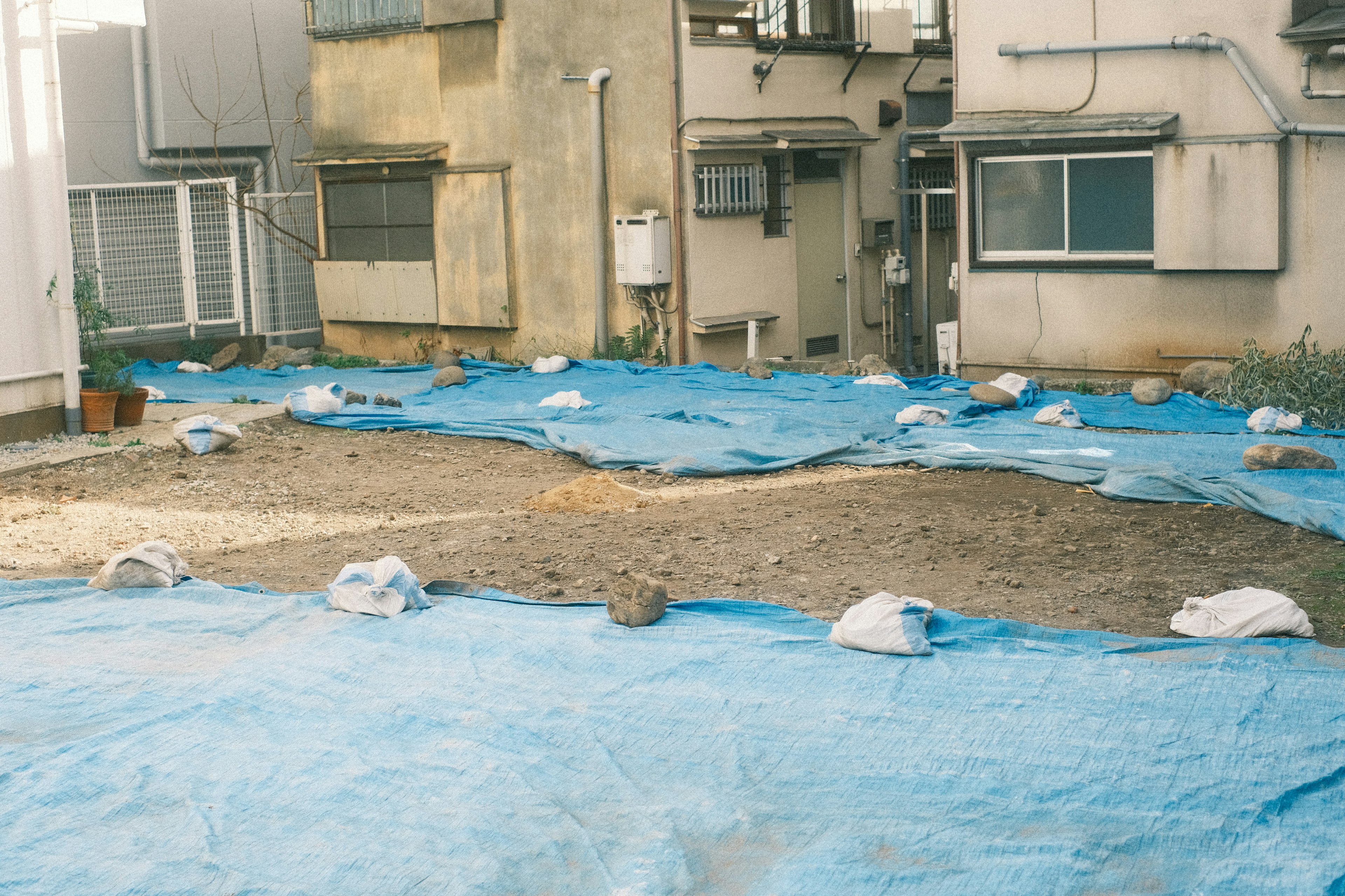 Ground covered with blue tarps in a backyard with surrounding buildings