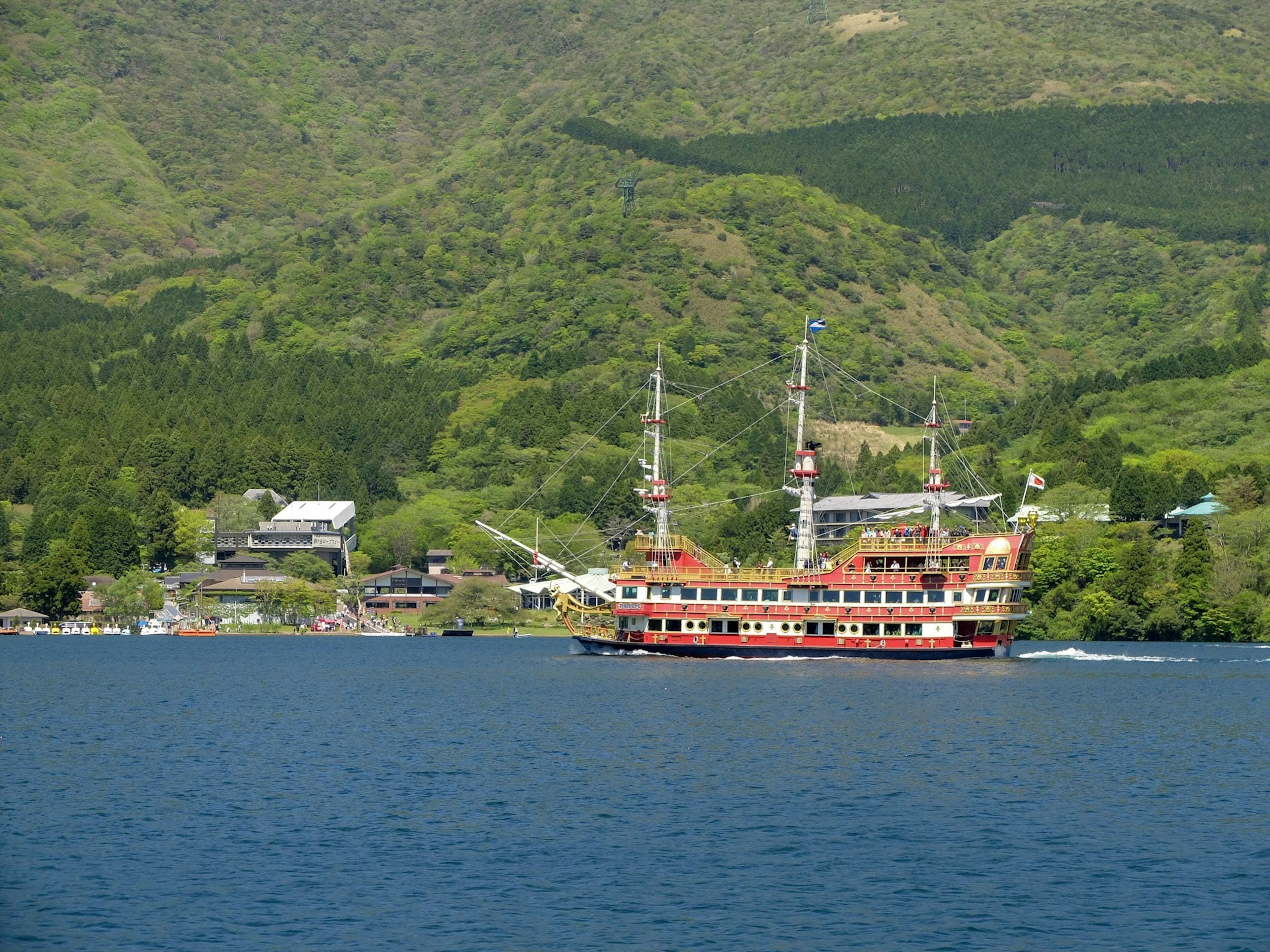 A red sailing ship navigating a lake with lush green mountains in the background