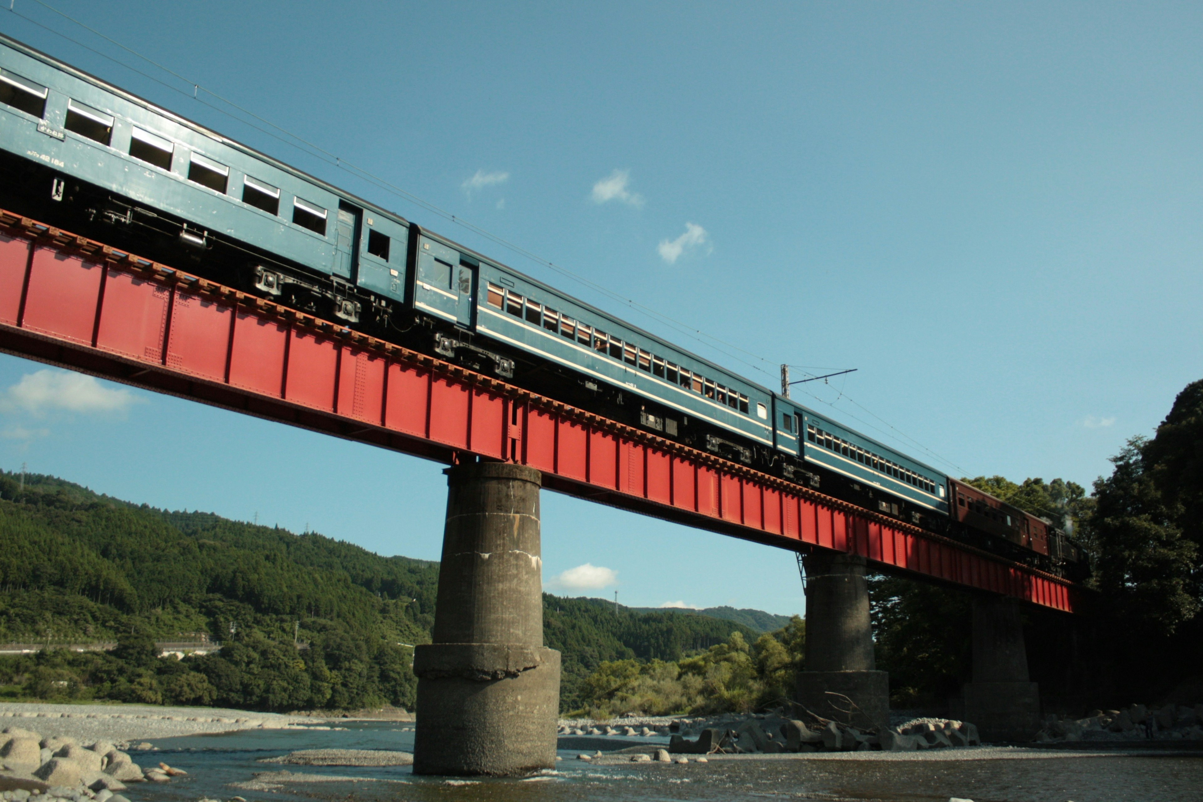 Kereta biru melintasi jembatan merah dengan latar belakang pemandangan indah