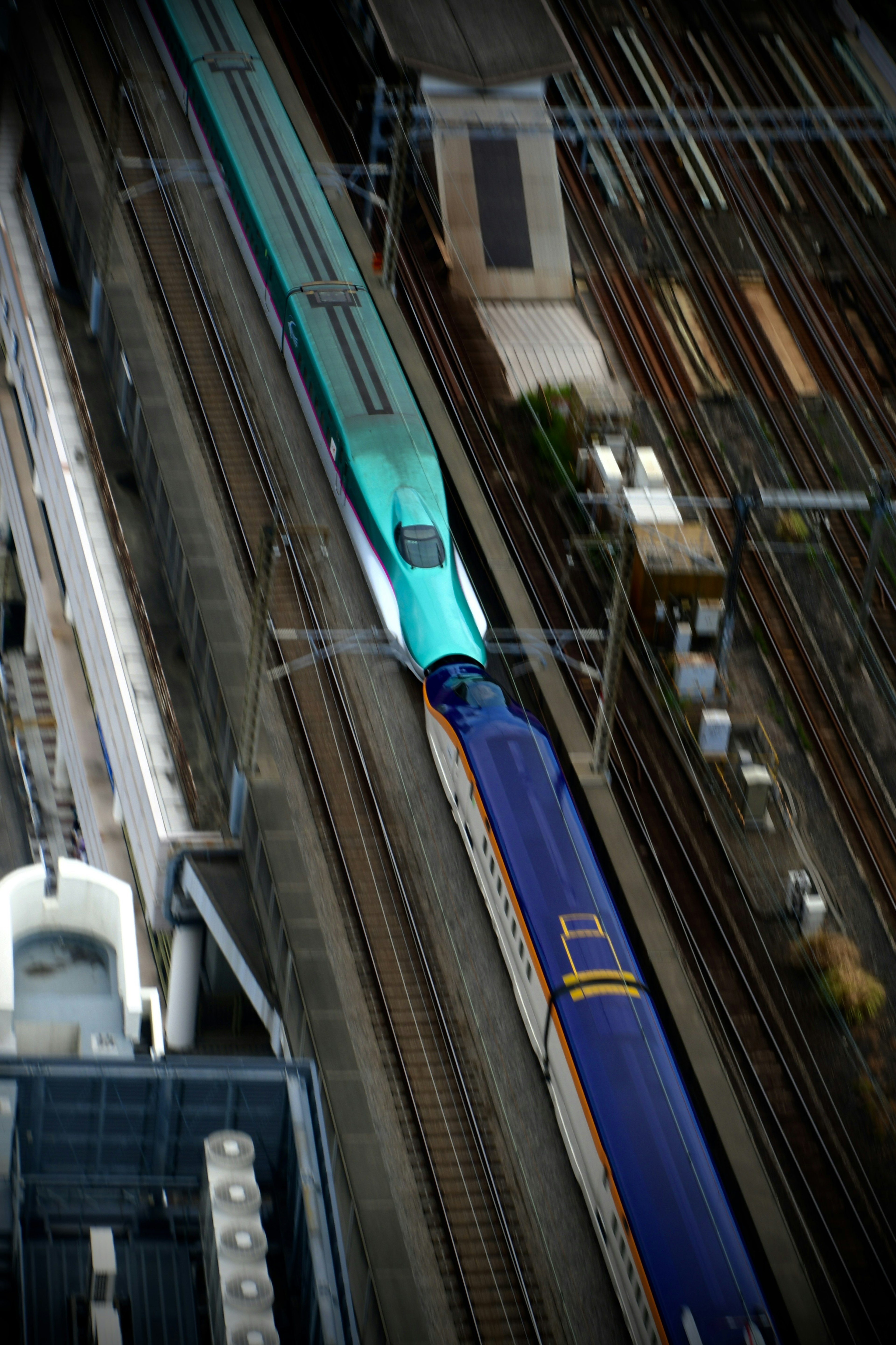 A blue and green Shinkansen train crossing tracks from an aerial view