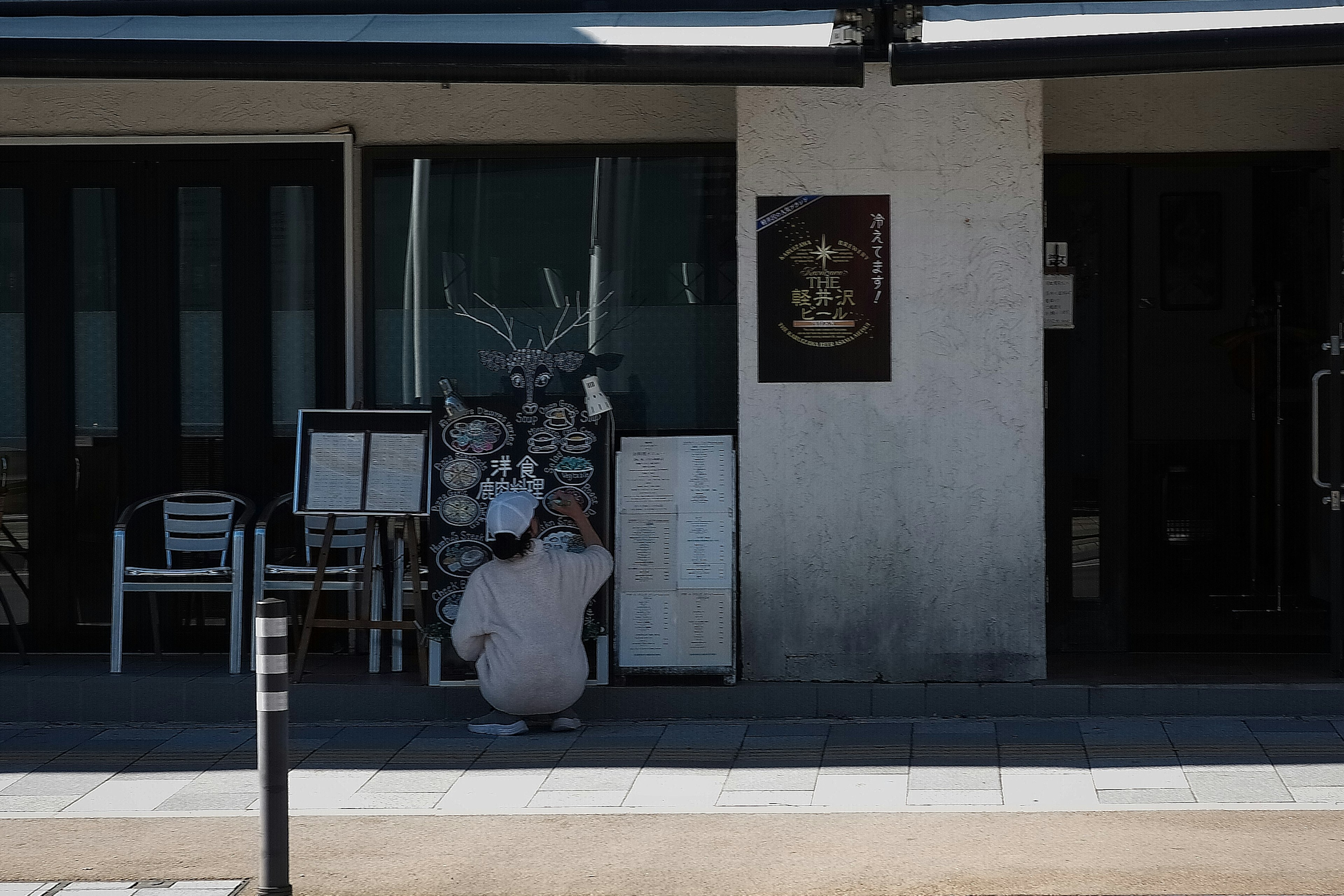 A person in white attire sits in front of a store with a menu board and decorative items in the background