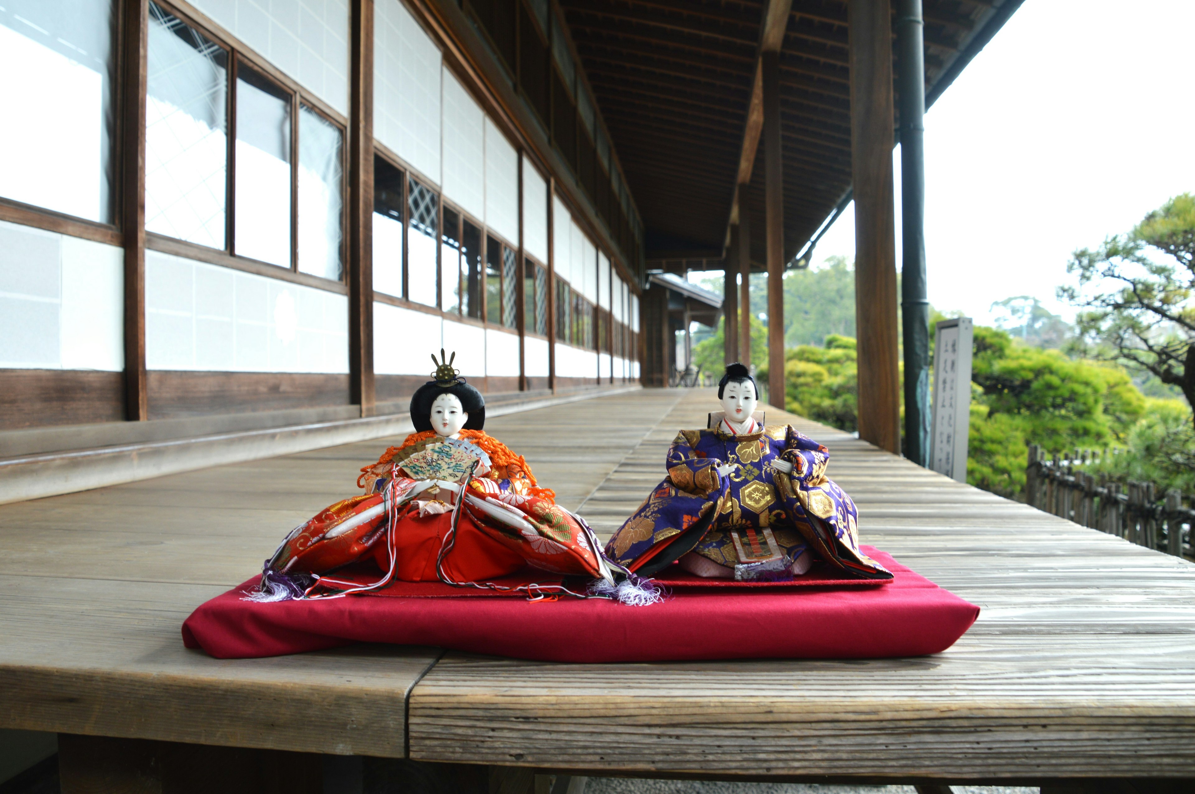 Poupées hina japonaises exposées sur une véranda en bois avec un bâtiment traditionnel et un jardin en arrière-plan