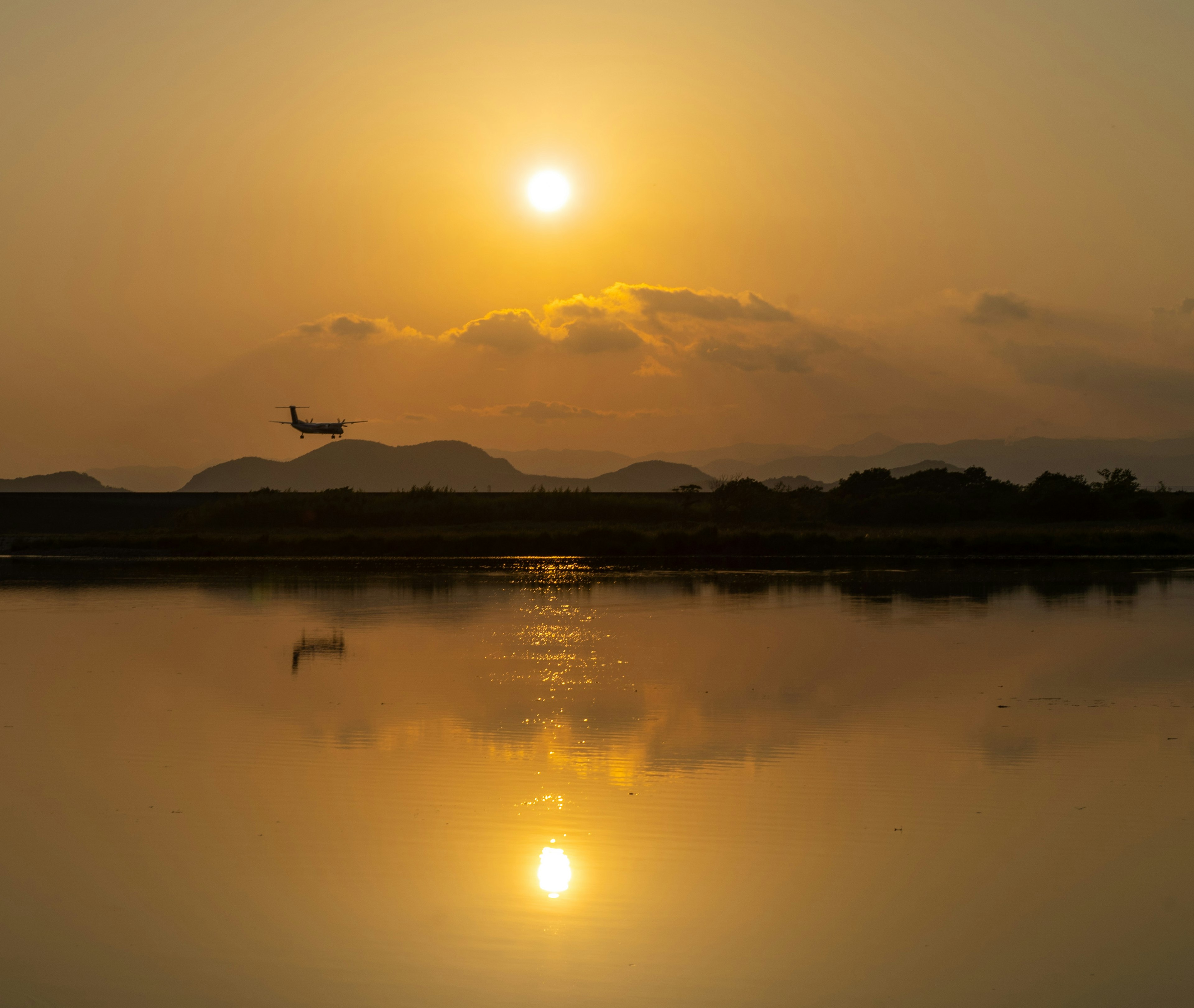 夕日が映る静かな湖面に飛行機が飛ぶ風景