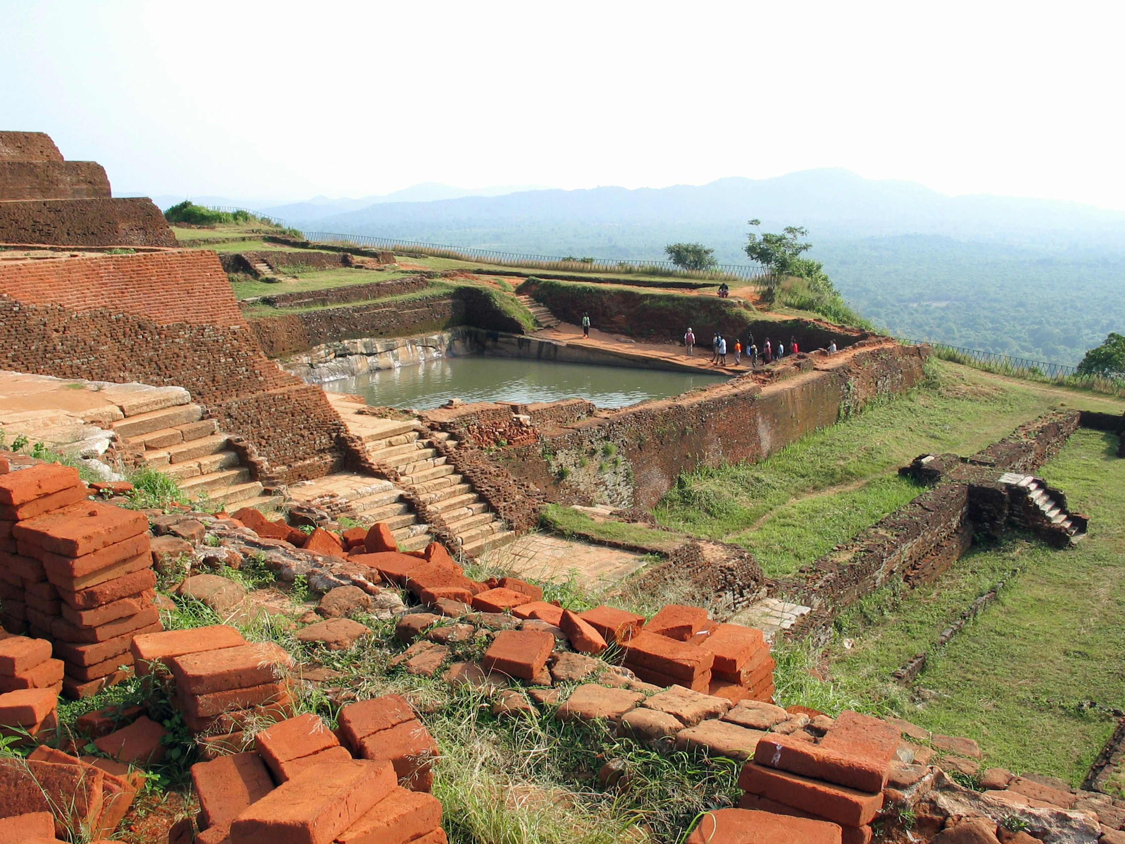 ทิวทัศน์ของซากโบราณสถานที่มีหญ้าสีเขียวและโครงสร้างอิฐแดง บันไดที่แตกหักและสระน้ำ