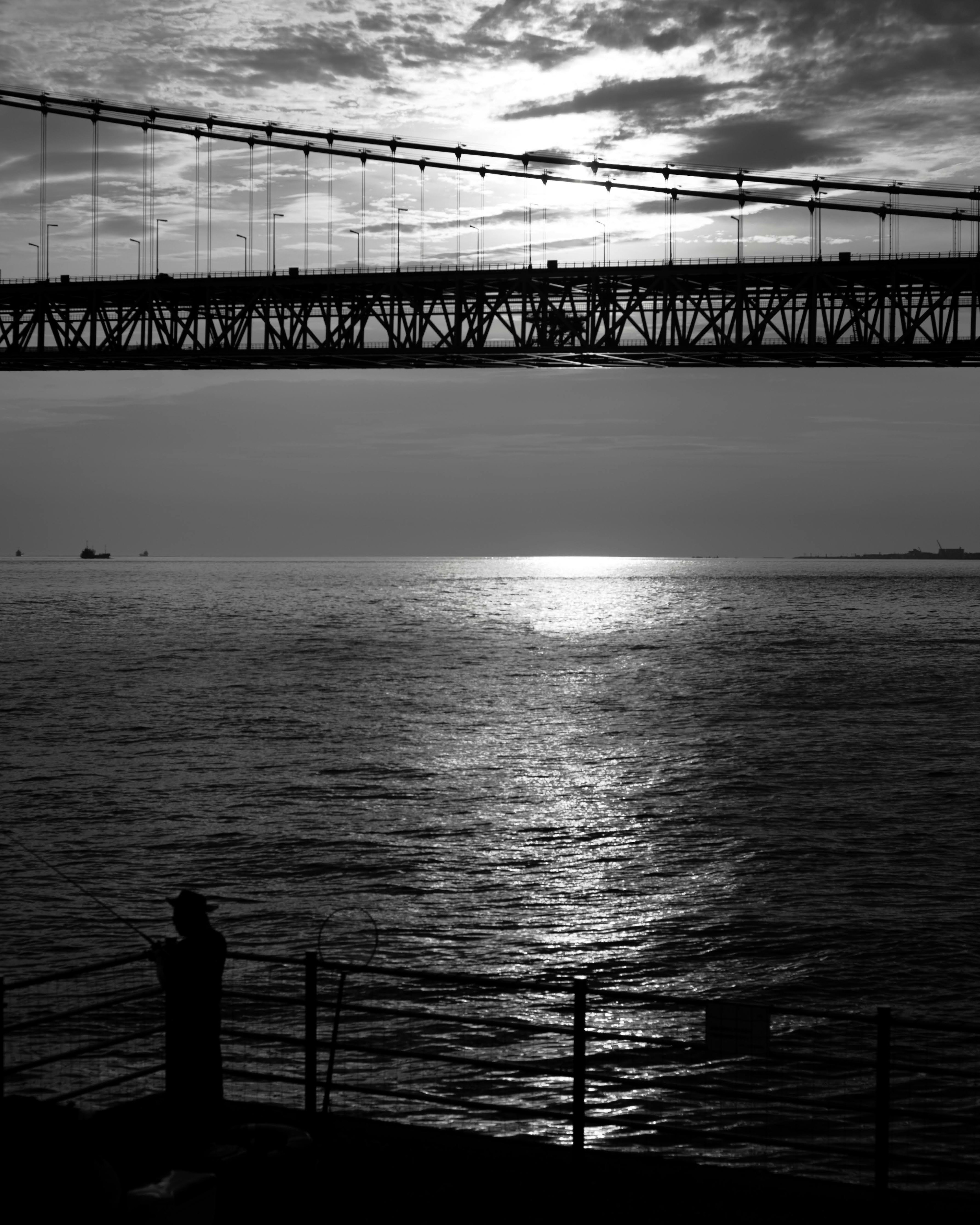 Pemandangan laut hitam putih dengan jembatan dan matahari terbenam yang memantul di air