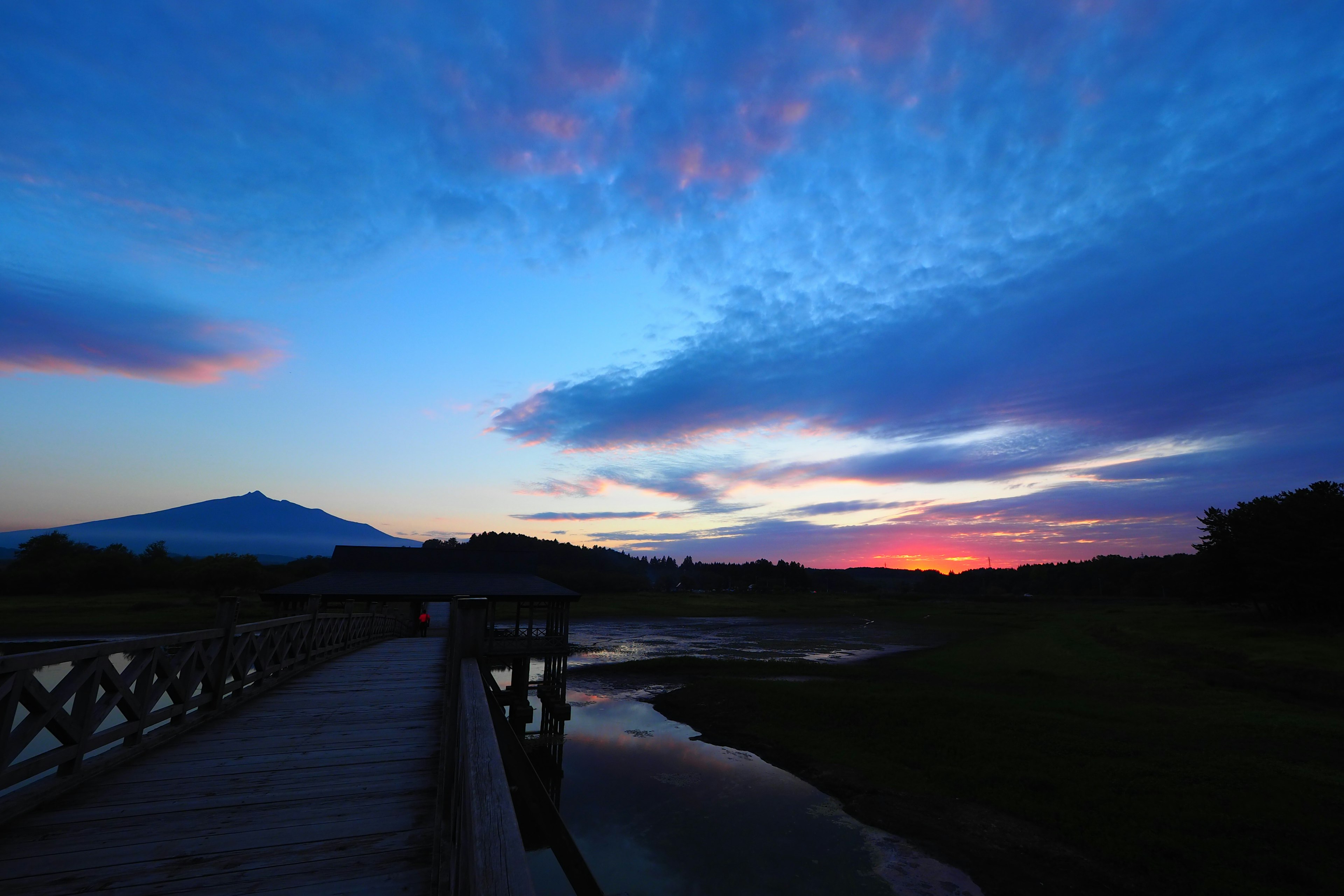 Vista escénica con un puente de madera bajo un cielo azul y tonos de atardecer y una montaña