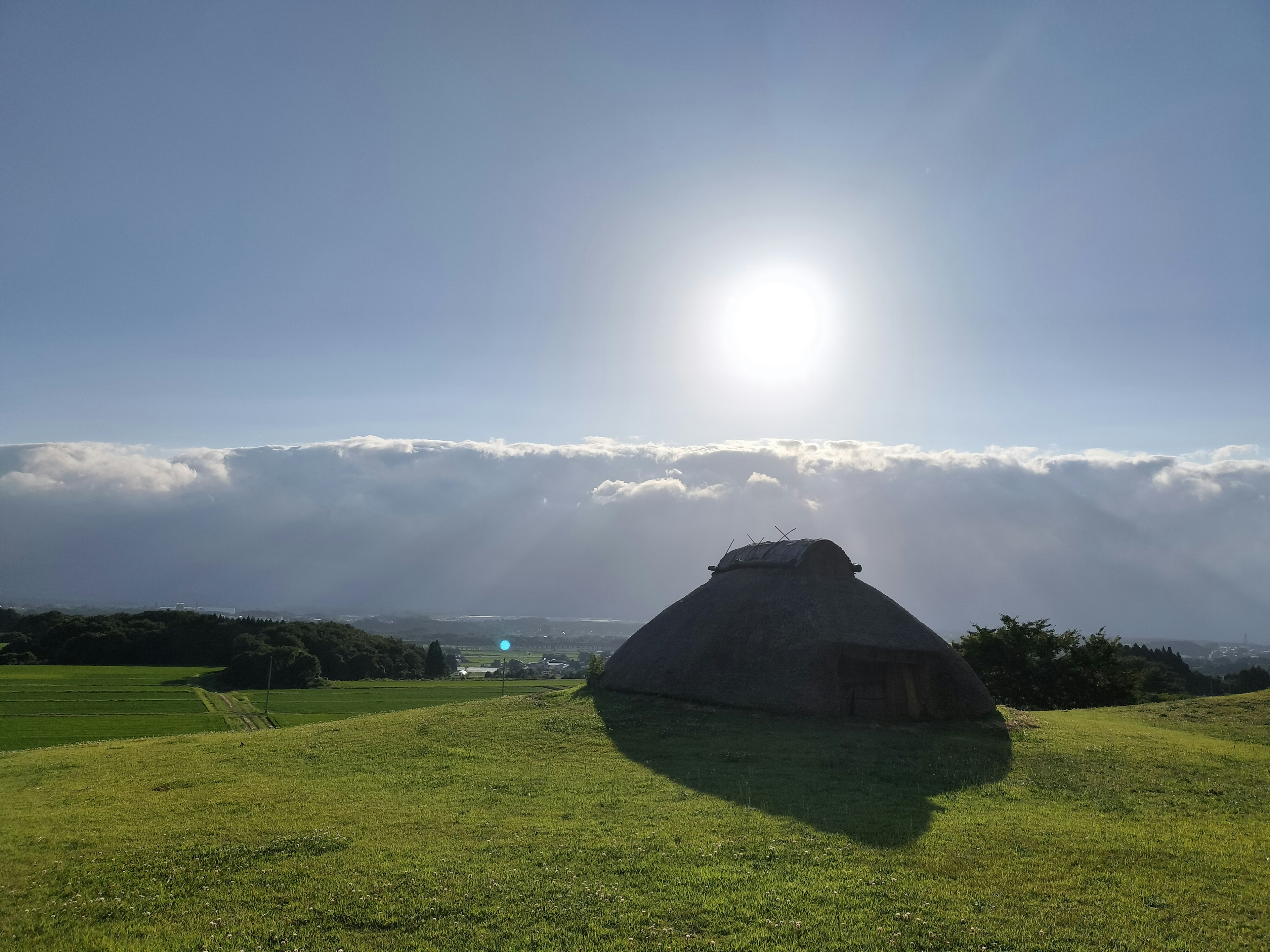 青空と太陽の下にある茅葺屋根の家と緑の草原